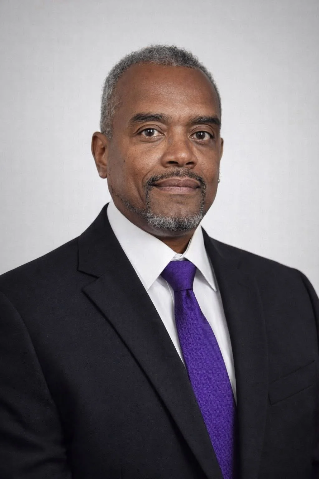 A professional man with gray hair and a beard, wearing a black suit, white shirt, and purple tie, standing against a plain background.