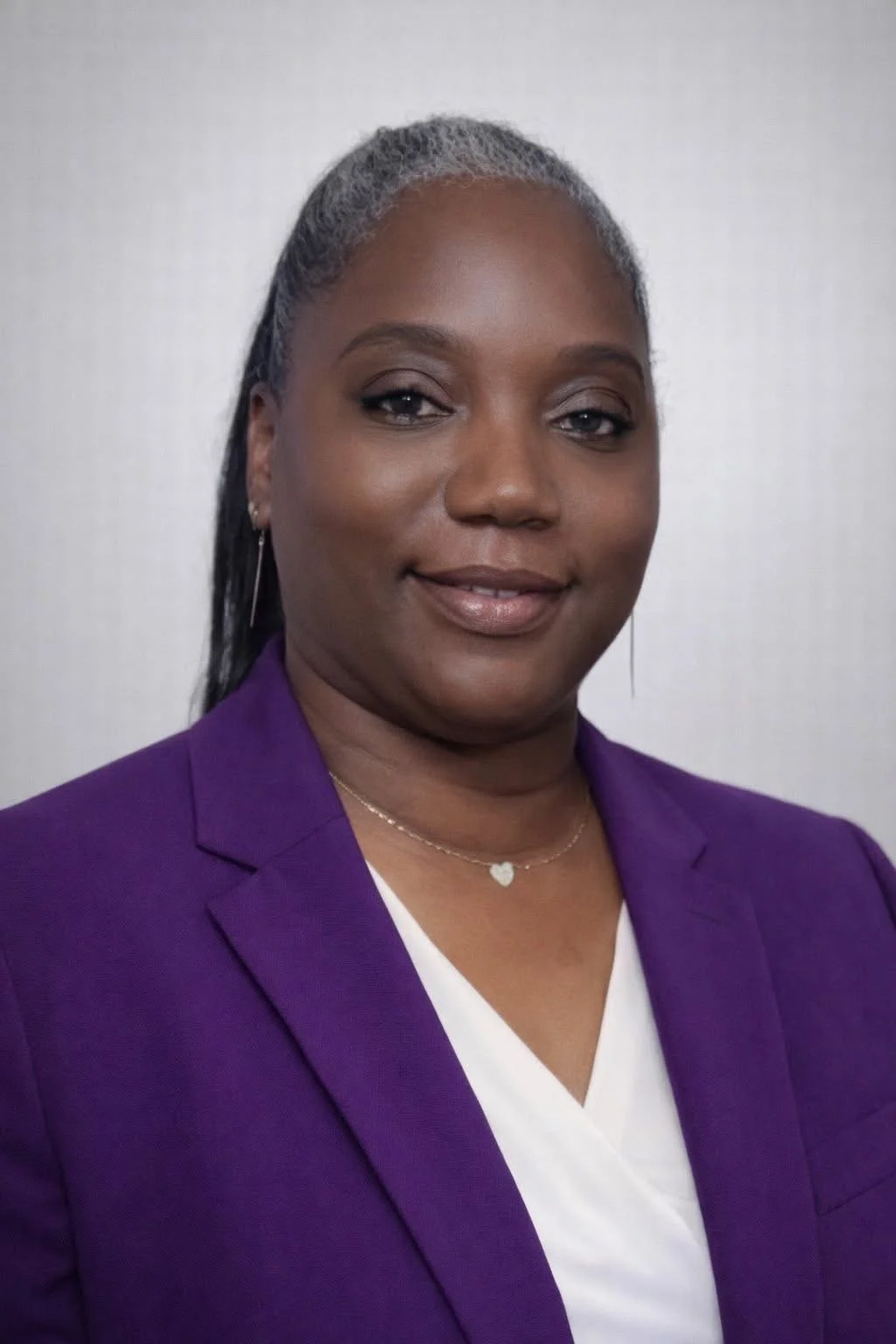 Portrait of an African American woman with gray hair, wearing a purple blazer, a white top, and a silver heart necklace, smiling softly.