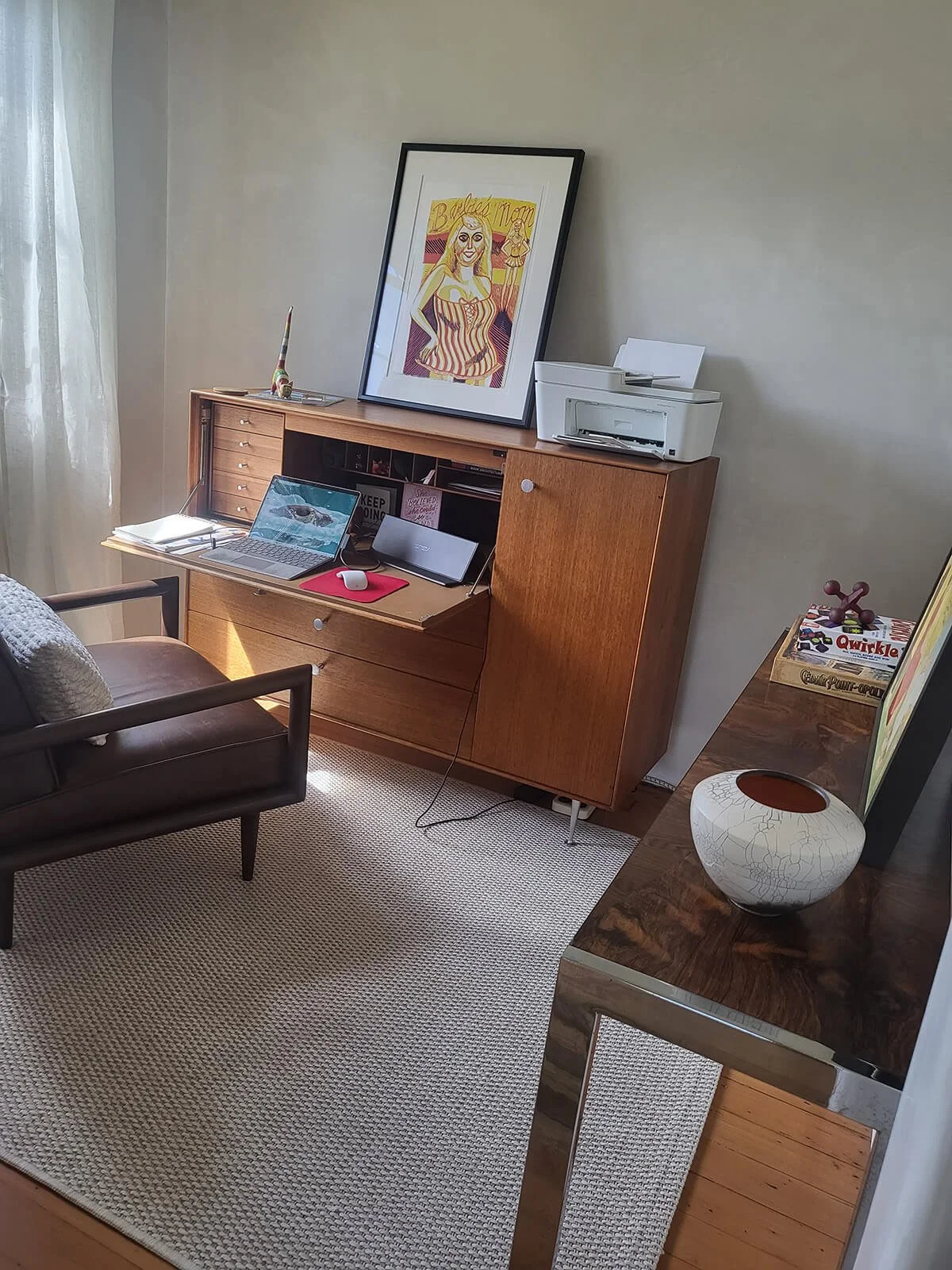 Home office with a wooden desk, laptop, printer, framed colorful artwork, and a black armchair with a cushion.