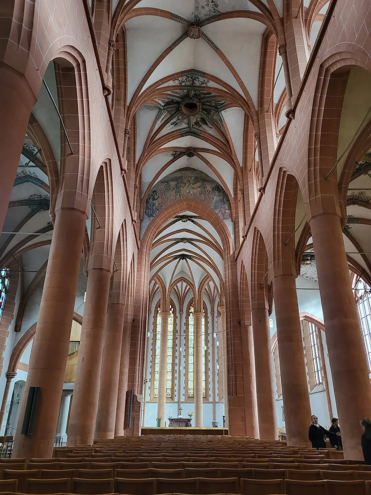 Interior of a Gothic-style church with tall pink stone columns, arched ceilings, and stained glass windows at the front.