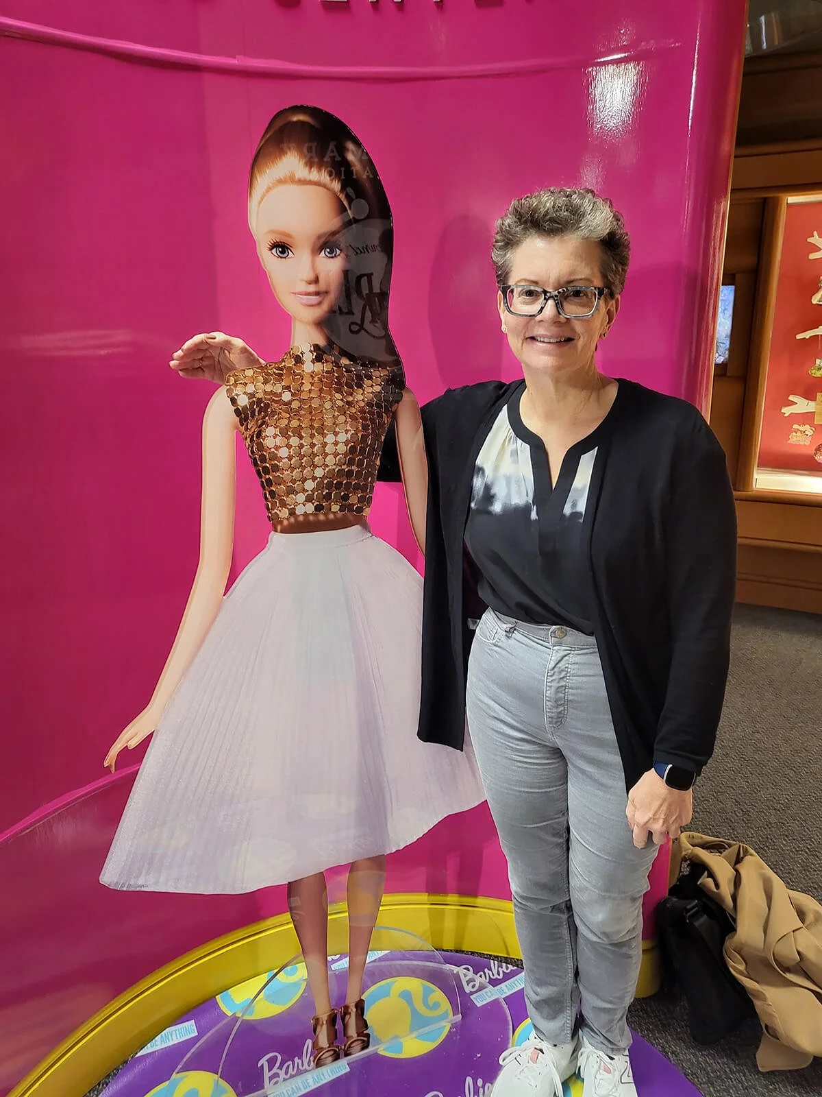 Woman standing next to a Barbie cutout, smiling, in a room with pink walls.