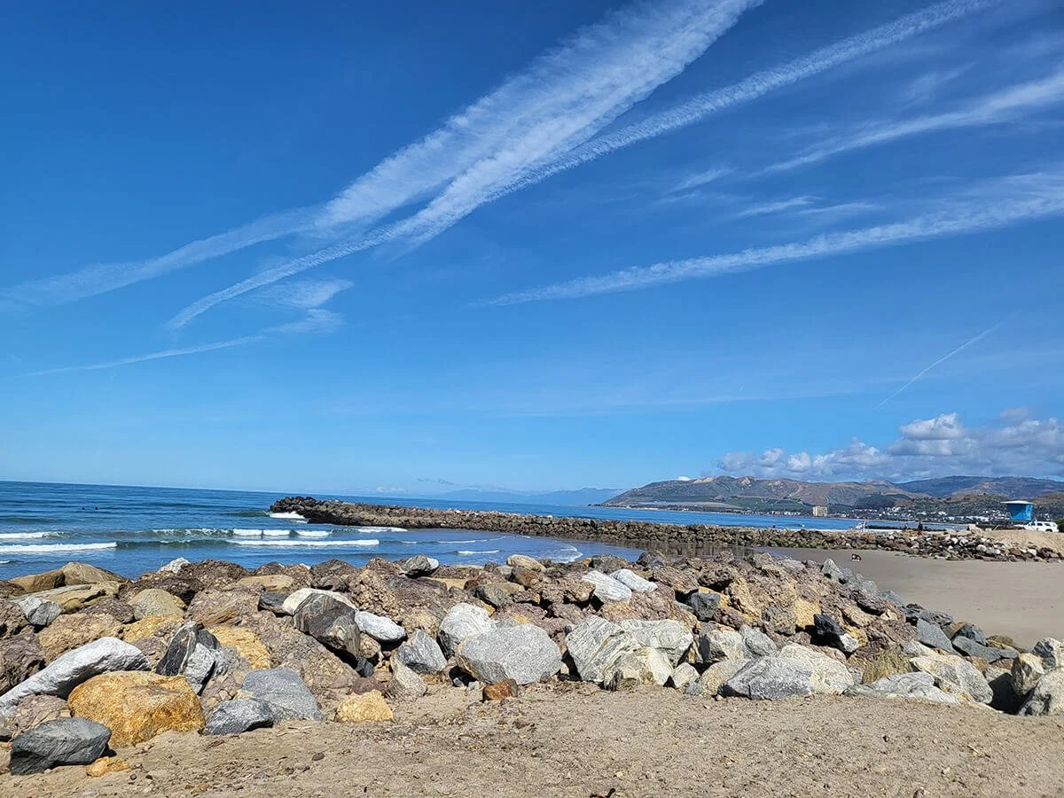 Sunny beach with rocks in the foreground, a rocky jetty extending into the ocean, and mountains in the distance under a blue sky with scattered clouds and contrails.