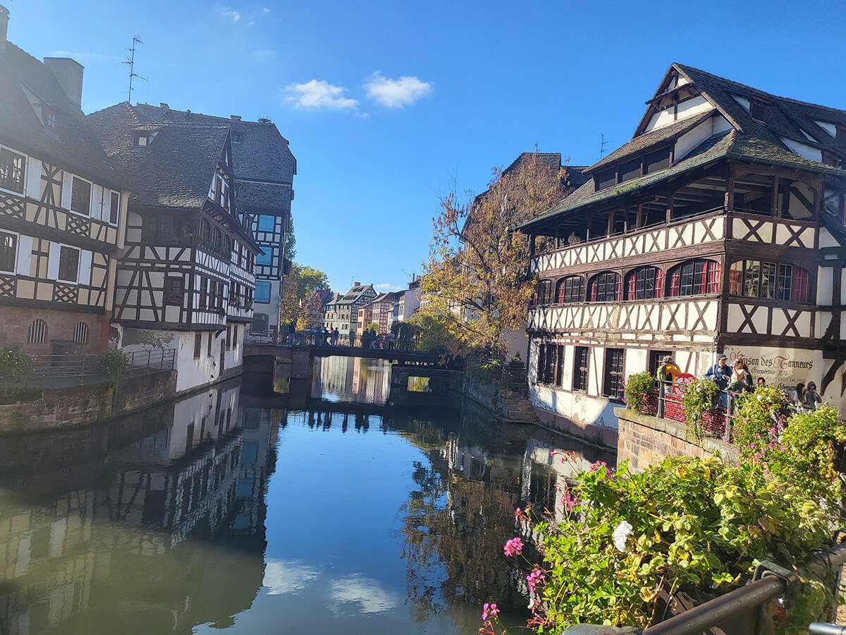 A picturesque view of a canal flanked by traditional half-timbered houses in a European town, with a small bridge, colorful flowers, and reflections in the water, under a bright blue sky.
