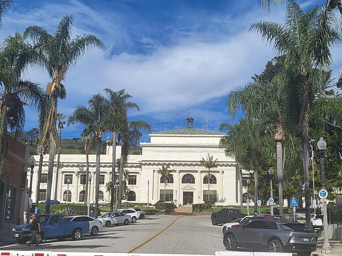 Historic white building with columns, surrounded by palm trees, in a parking lot filled with cars, under a partly cloudy sky.