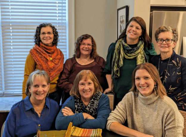 Seven women gathered indoors, smiling for a photo, with a window and kitchen in the background.