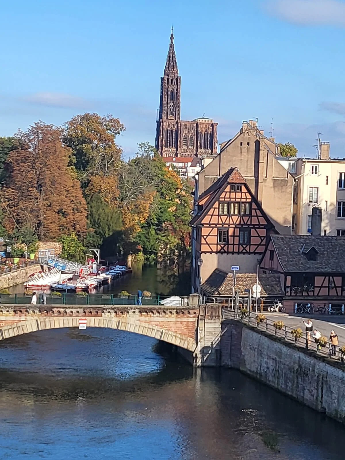 Scenic view of a small river with boats docked along the bank, a stone bridge in the foreground, and traditional European-style buildings with timber framing. A large Gothic church spire rises in the background under a blue sky with some clouds.
