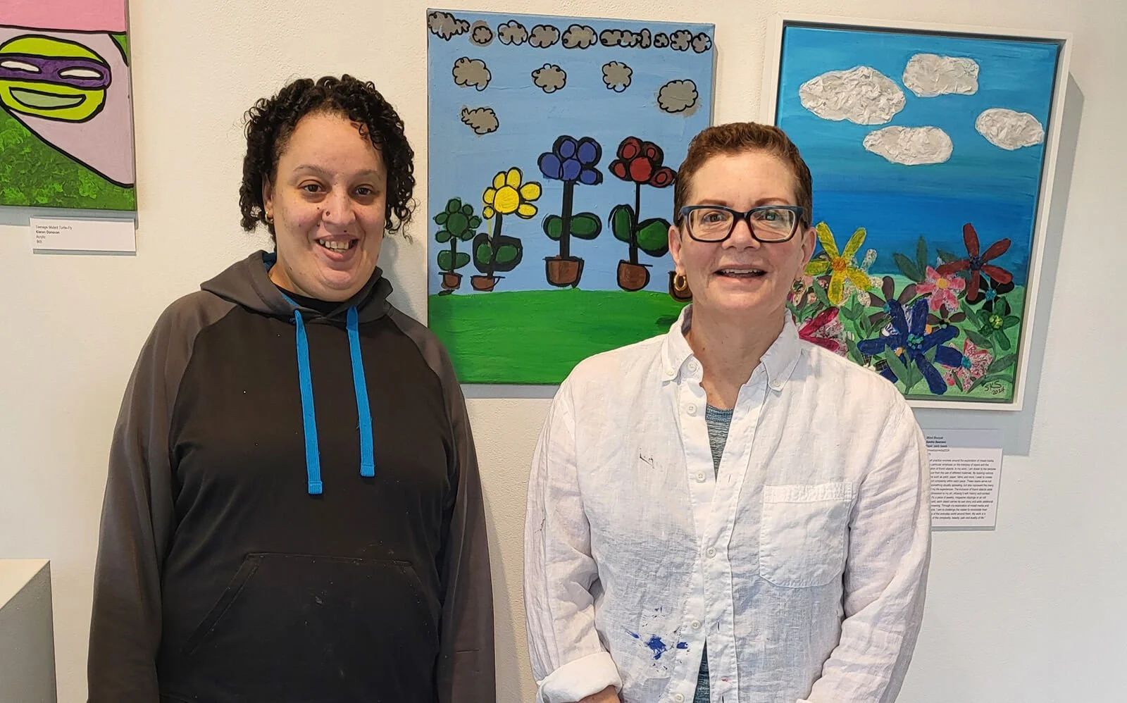 Two women standing in front of colorful paintings of flowers and clouds displayed on a gallery wall.