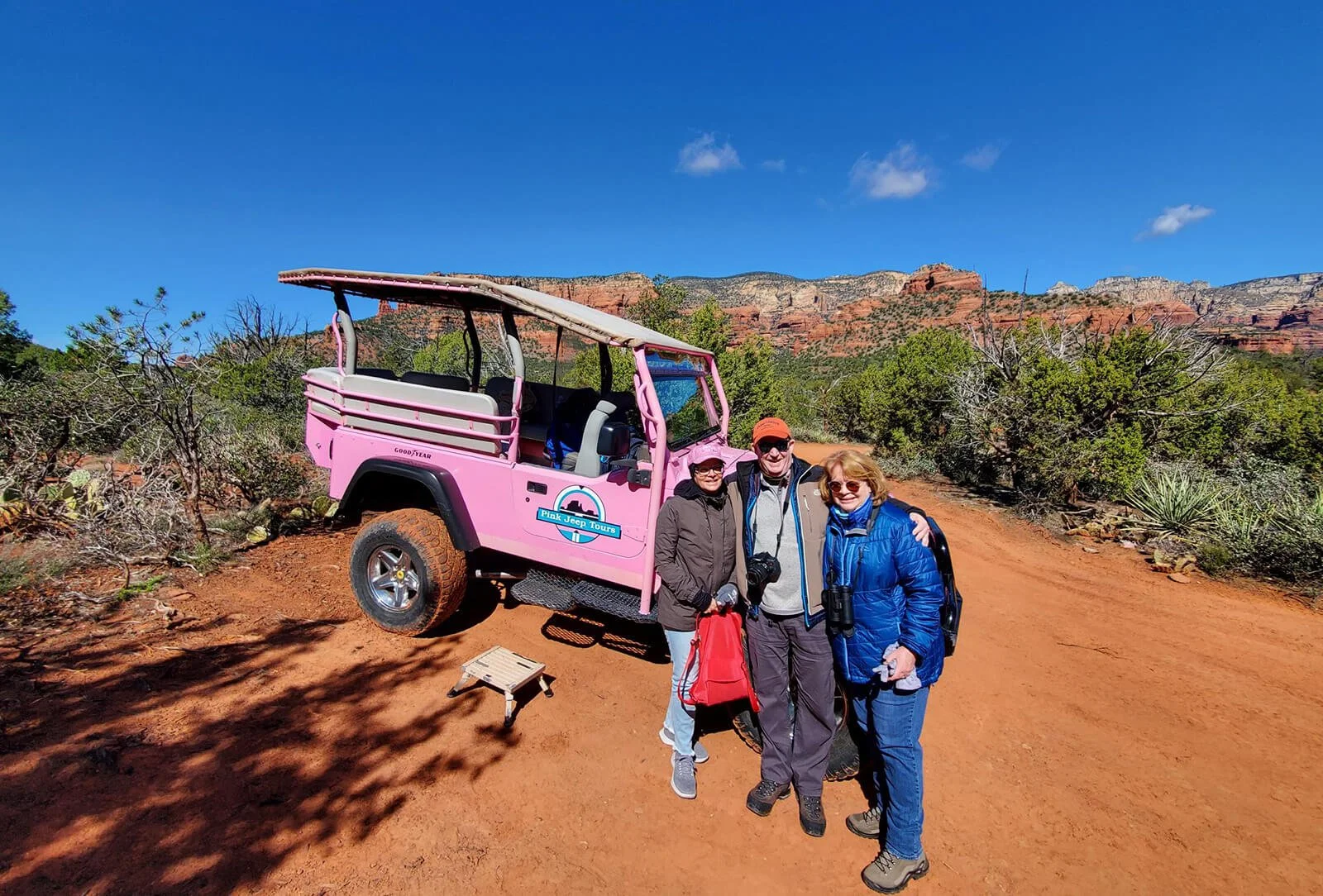 Three people standing together in front of a pink jeep with tour signage, in a desert landscape with red rocks and green bushes under a blue sky.