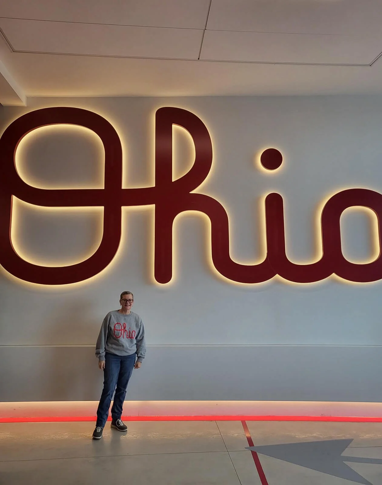 A woman standing in front of a large Ohio sign with backlit letters, wearing a gray Ohio sweatshirt and jeans, smiling.