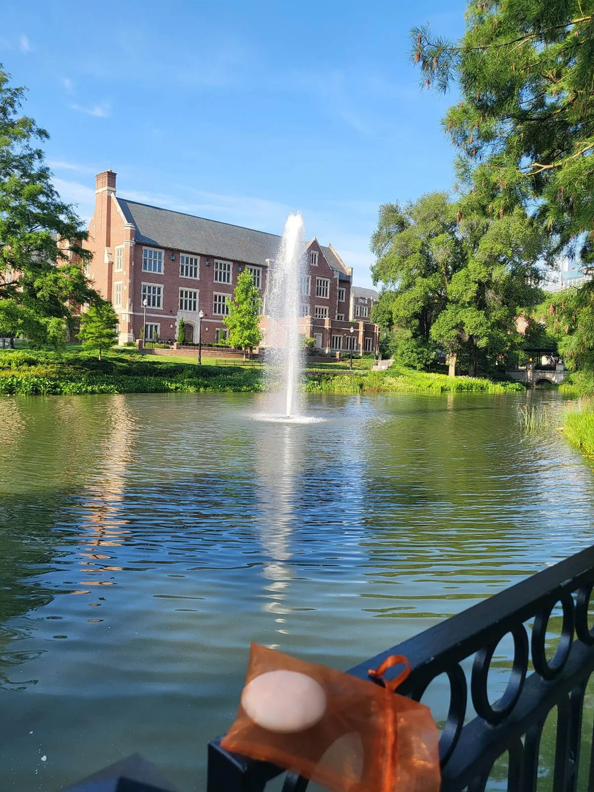 A pond with a fountain in the center, surrounded by trees and a brick building in the background, under a clear blue sky.