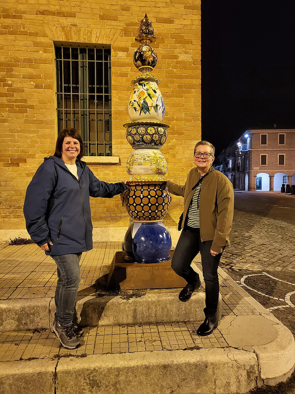 Two women standing on a sidewalk at night, smiling and posing with a colorful ceramic sculpture of stacked, decorated bowls and vases against a brick building with barred window.