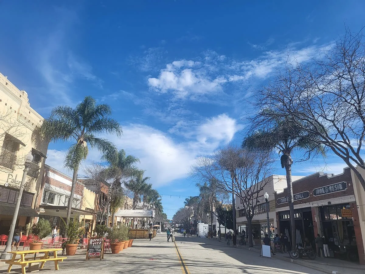 A pedestrian street with shops, palm trees, and people walking under a blue sky with scattered clouds.