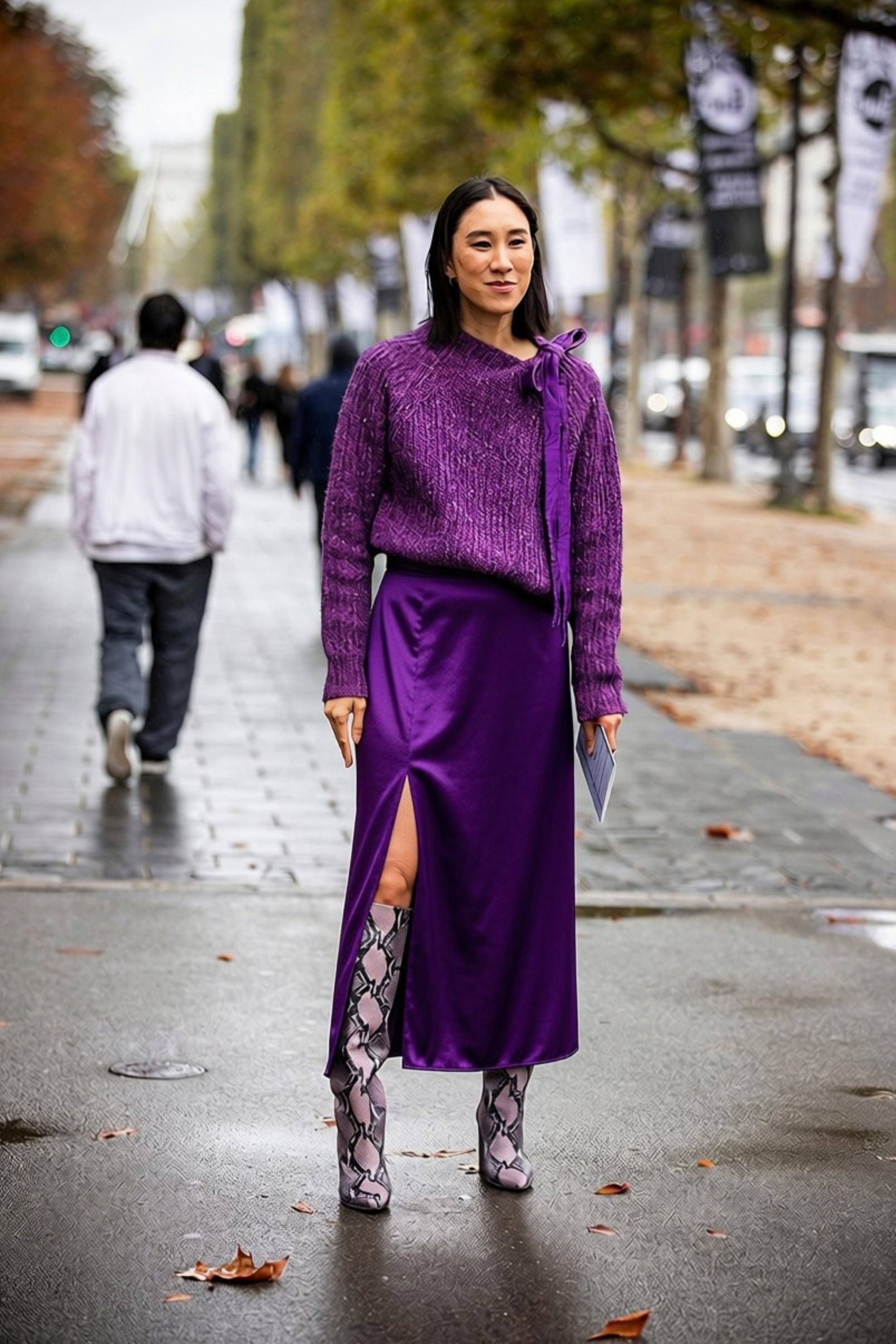 woman walking outdoors on a city street with fallen leaves, wearing a purple outfit with patterned thigh-high boots.