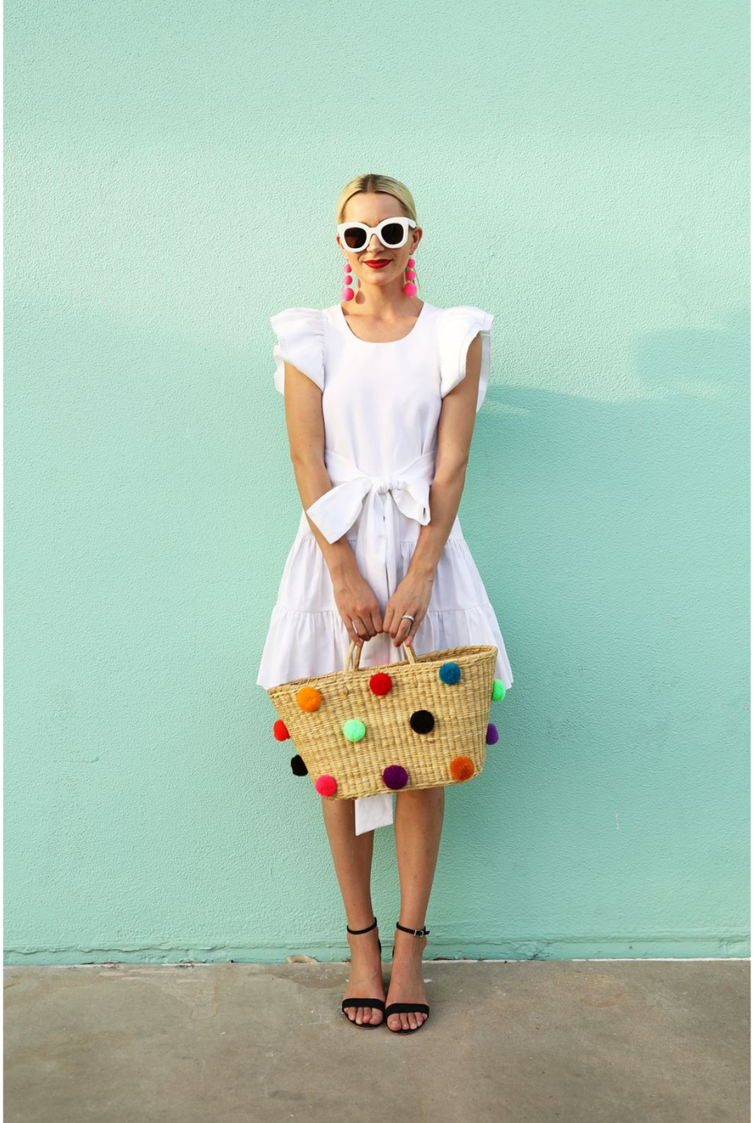 women in a white dress against a green wall bolding a Nannacay basket with pompoms