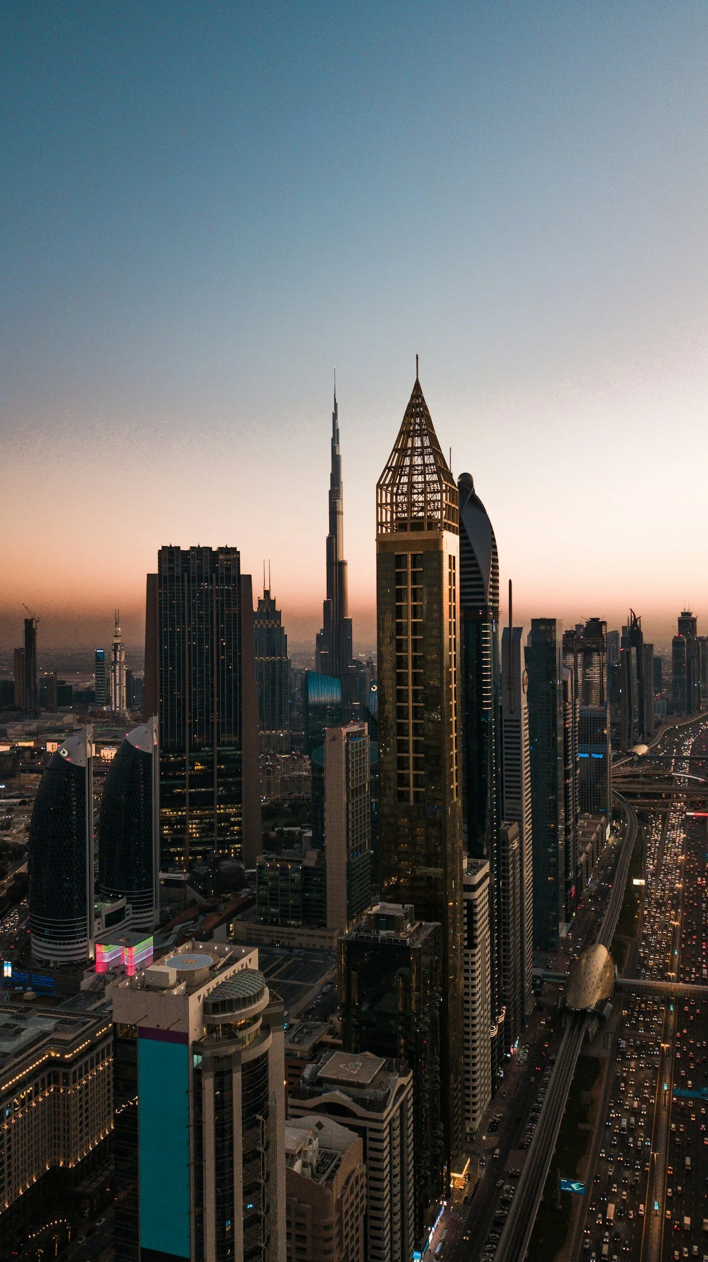 A city skyline at dusk with tall skyscrapers, including the Burj Khalifa, Dubai's tallest building, with a clear sky and some city lights.