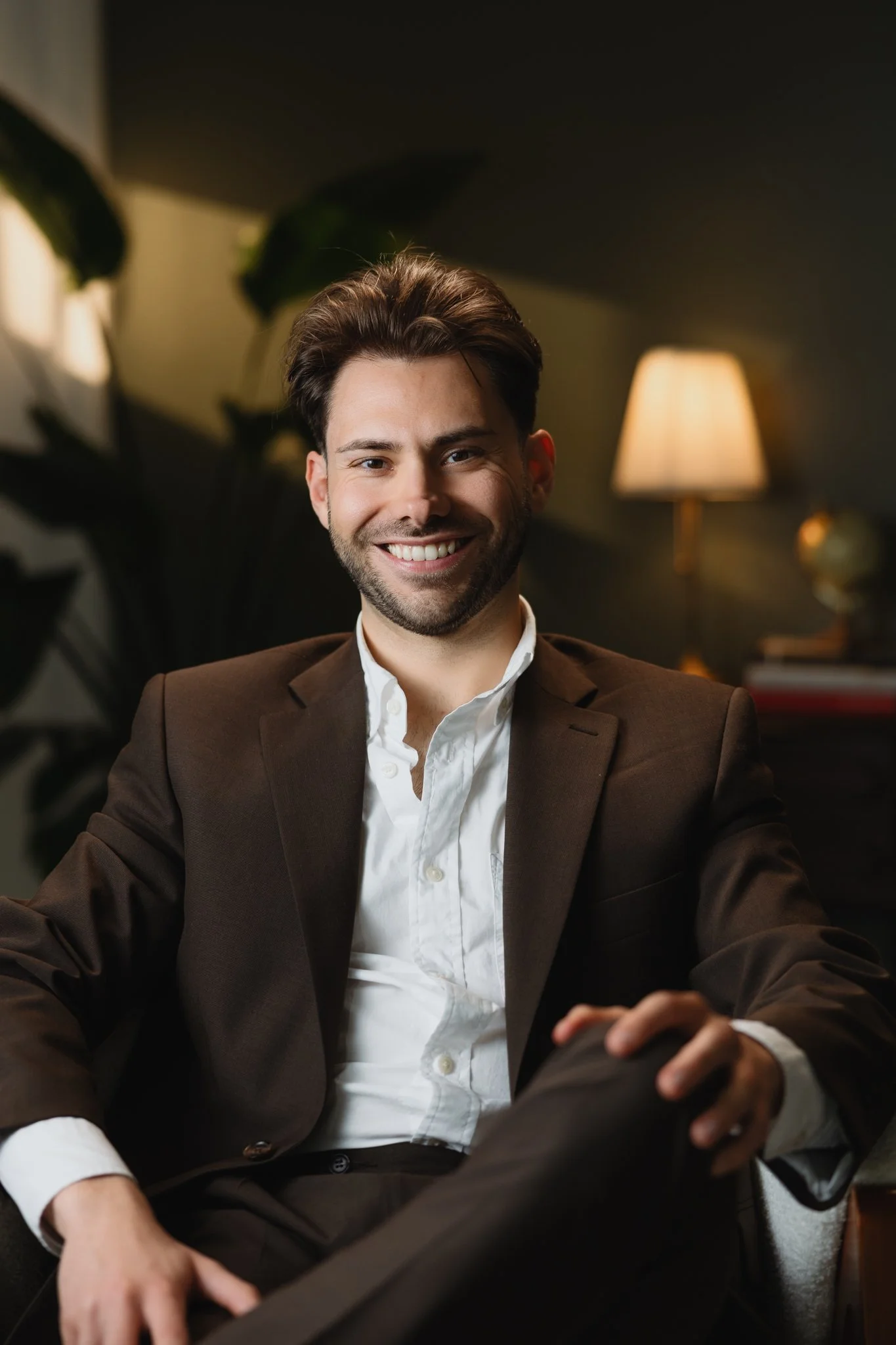 A young man with dark hair and a beard, smiling, dressed in a brown suit and white shirt, sitting in a cozy indoor setting with warm lighting, plants, and lamps in the background.