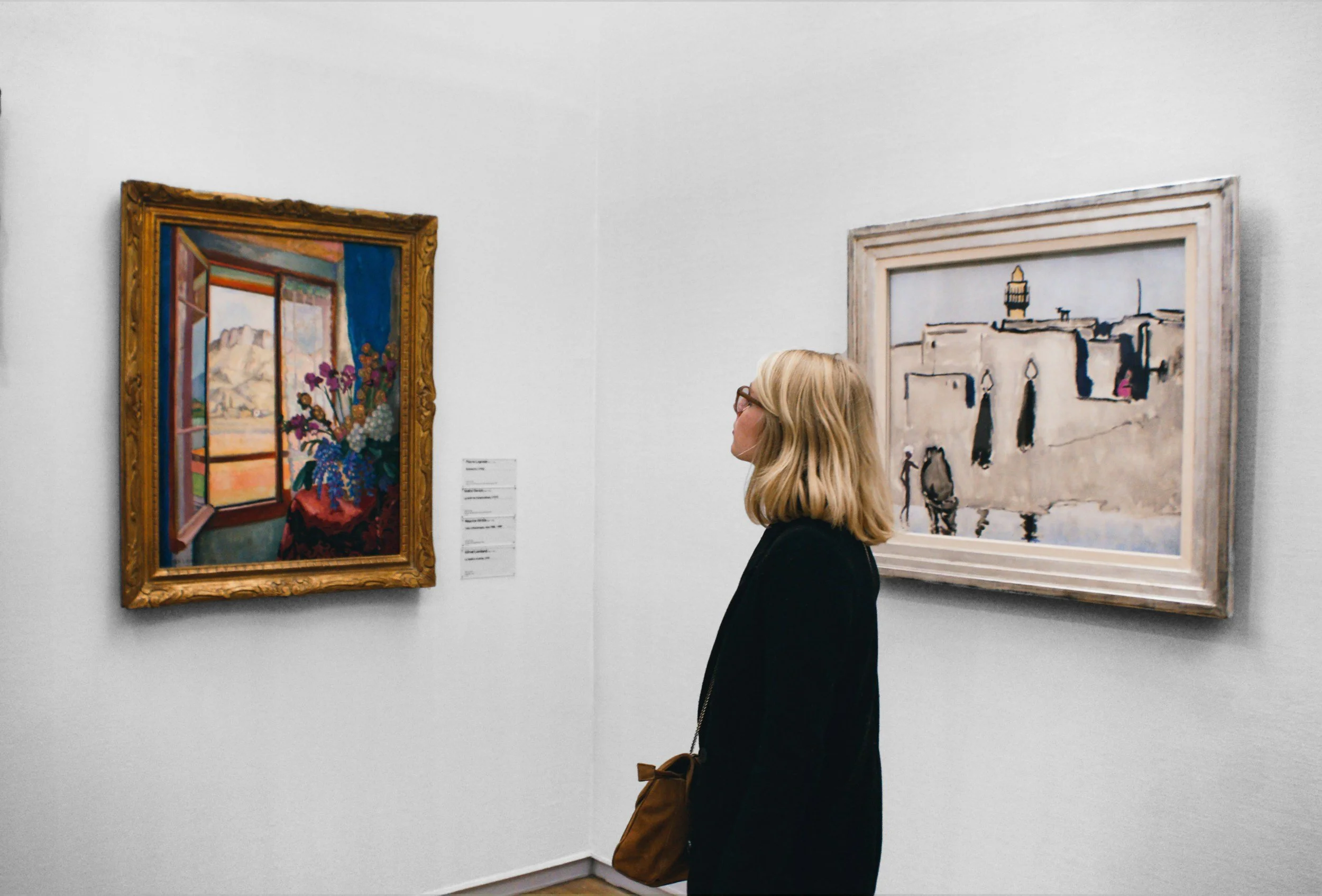 A woman with blonde hair, glasses, wearing a black coat, and carrying a brown bag, standing in front of two paintings in an art gallery.