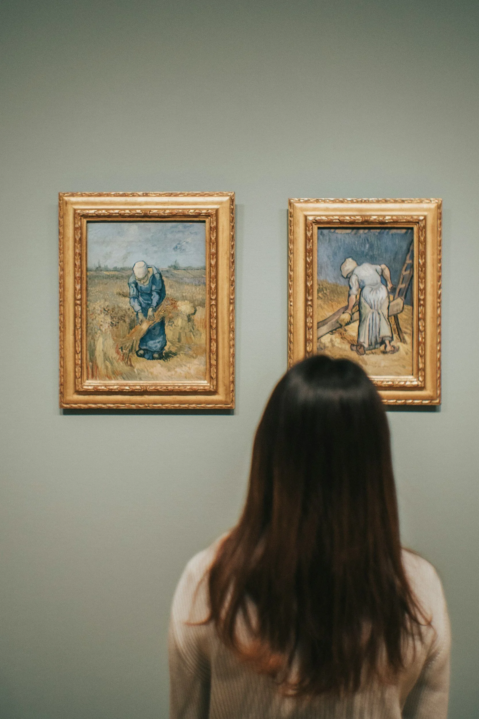 A woman with brown hair looking at two paintings of women working in fields displayed on a gallery wall.
