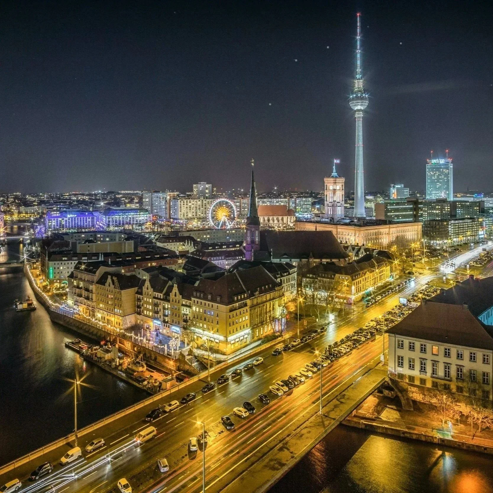 Nighttime view of a city skyline featuring a tall telecommunications tower, a Ferris wheel, historic buildings, and illuminated streets along a river.