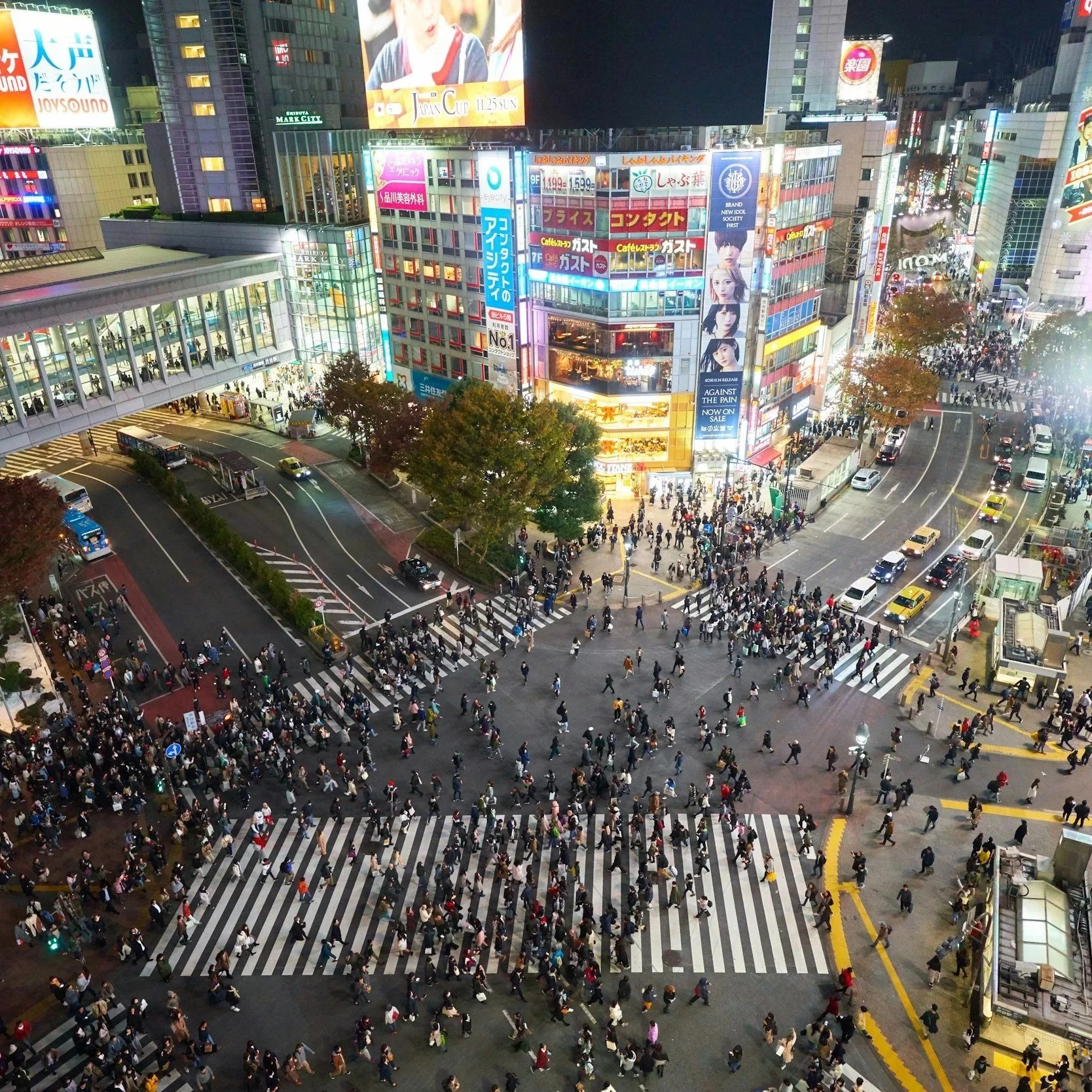 Nighttime aerial view of a busy city intersection filled with pedestrians crossing crosswalks, lined with tall illuminated buildings with colorful advertisements and billboards.