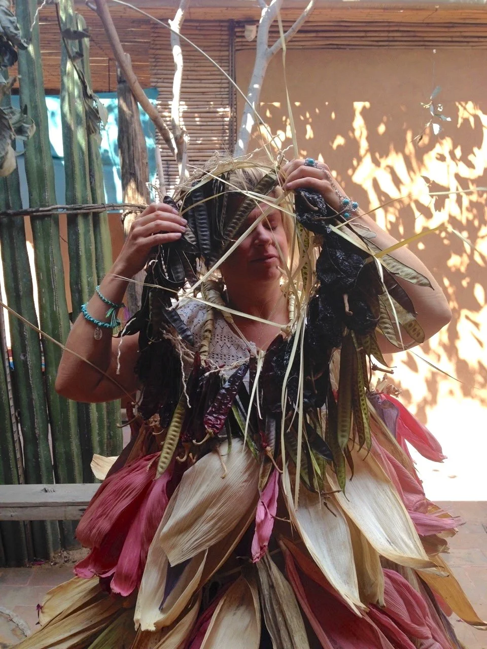 Woman wearing traditional clothing made of large, colorful dried leaves and feathers, with her hands on her head, standing outdoors surrounded by bamboo plants and sunlight.