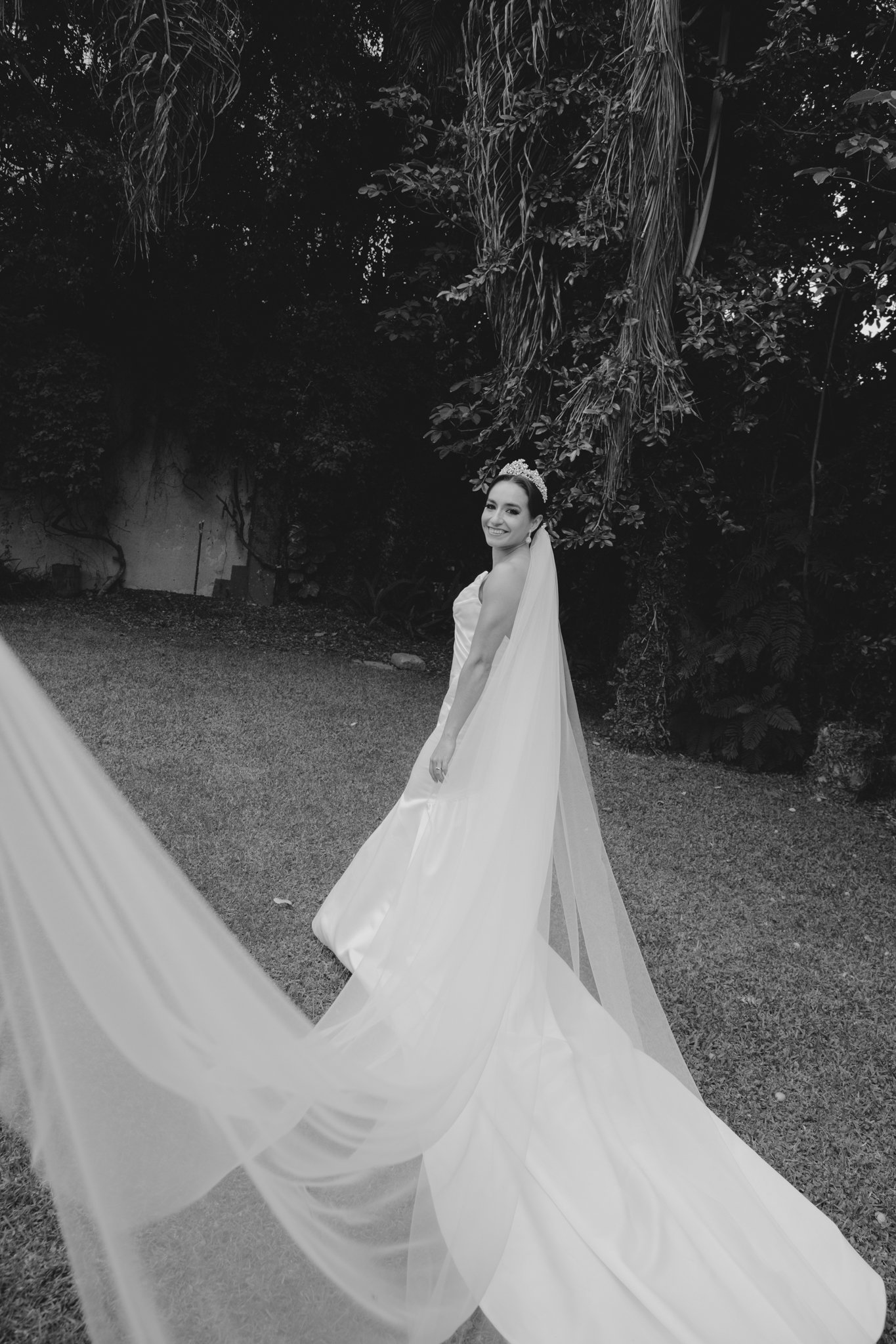 Una novia con vestido de boda, diadema y velo, sonriendo en un jardín con árboles y vegetación.