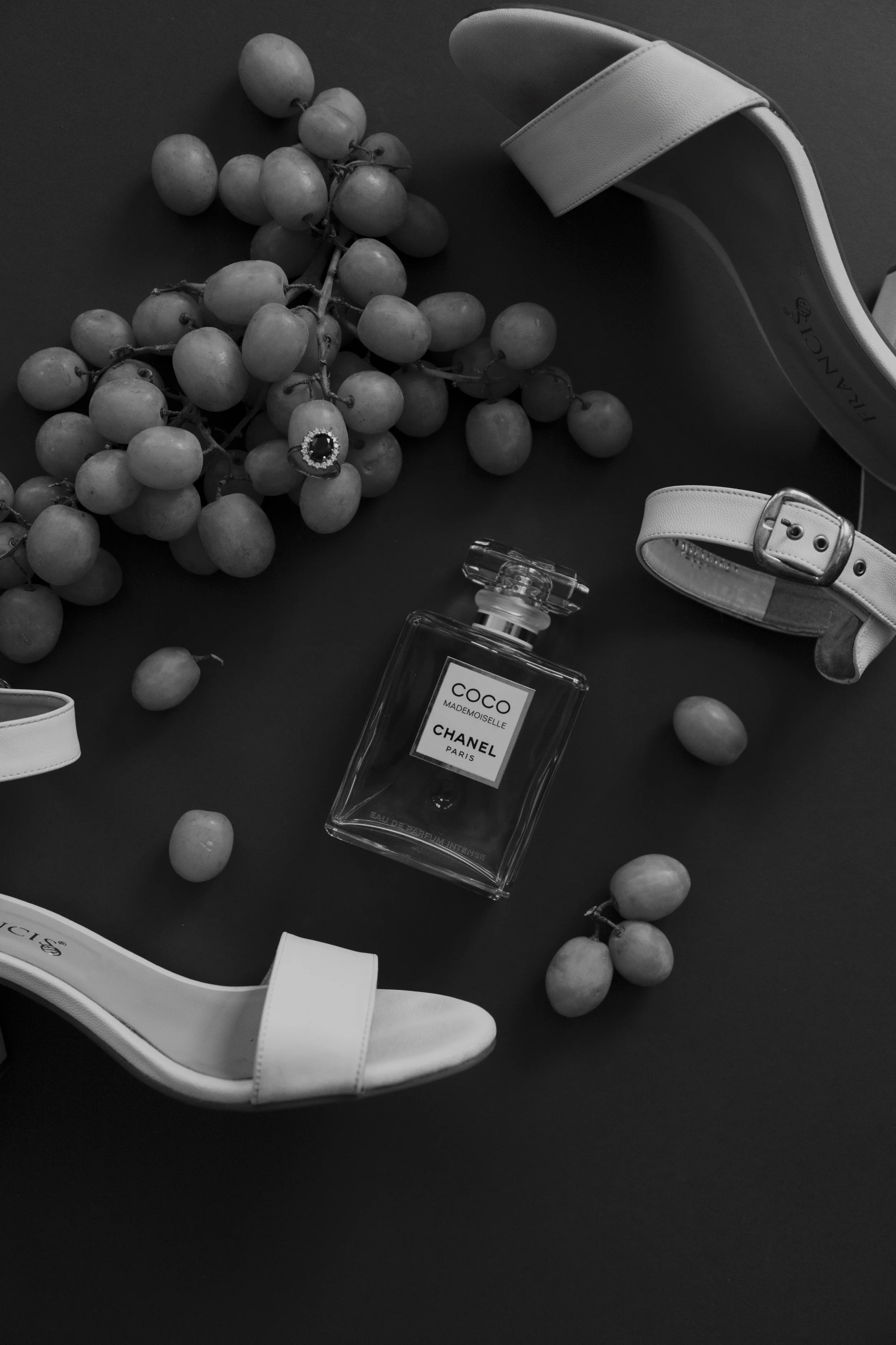 Ramo de uvas, perfume Chanel Coco repentino, zapatillas blancas de novia, y accesorios en un fondo oscuro.