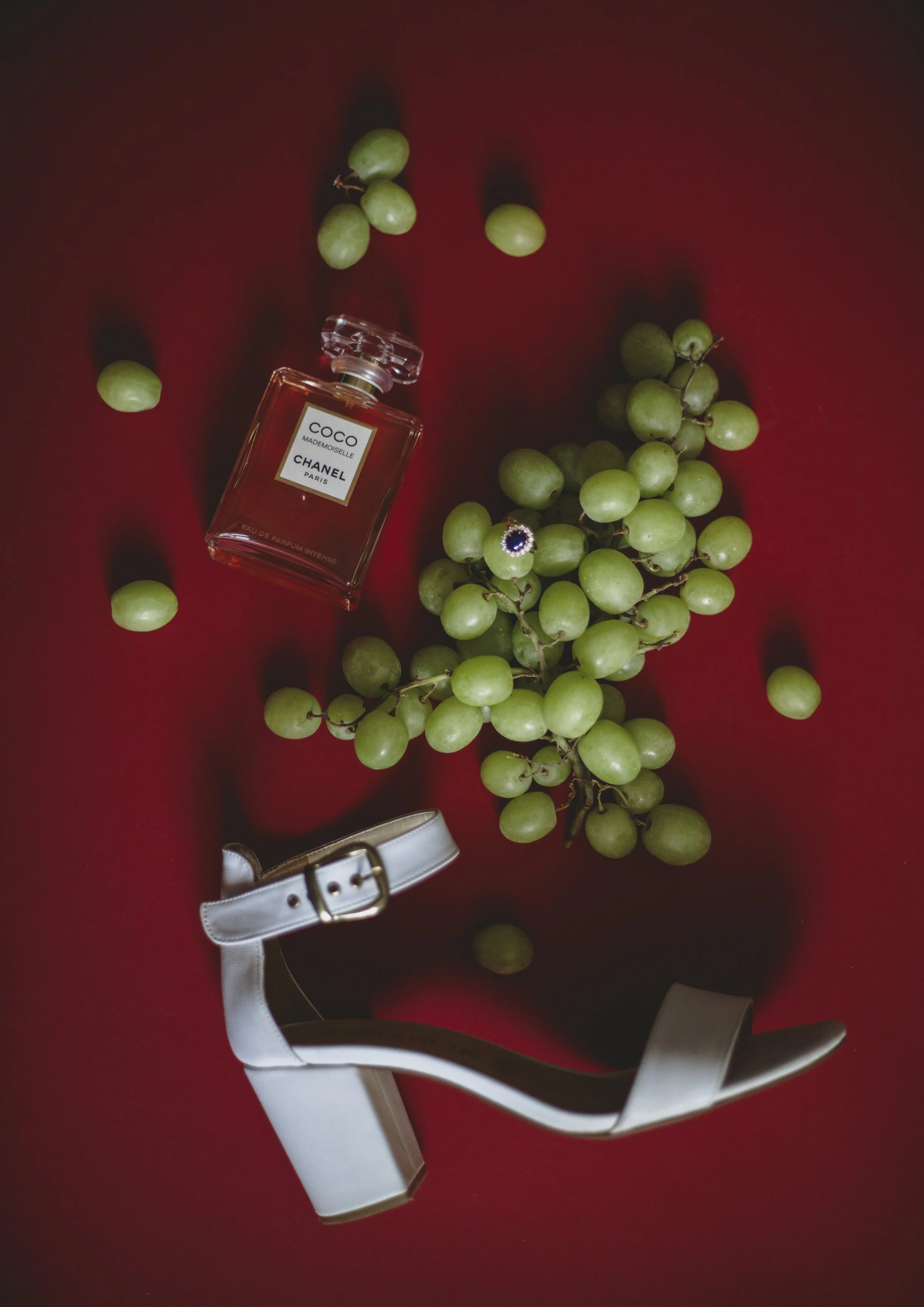 Botella de perfume Chanel Coco Mademoiselle, racimo de uvas verdes, una pulsera con piedra azul y oro, y un zapato de tacón grueso blanco sobre fondo rojo.