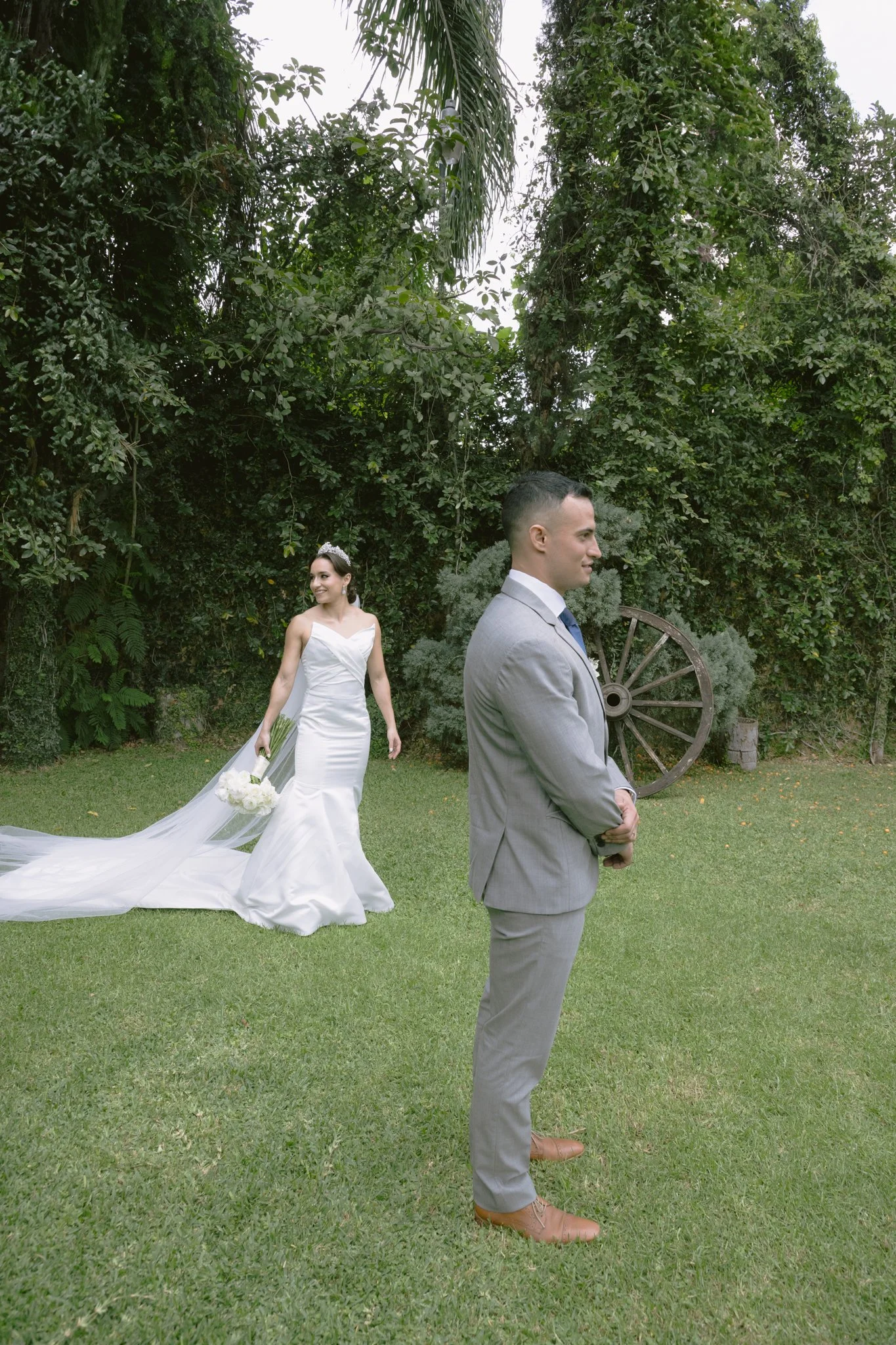 Una pareja de novios en una boda al aire libre. La novia lleva un vestido blanco largo con velo, sosteniendo un ramo de flores blancas. El novio, de espaldas y en primer plano, viste un traje gris y está sonriendo, mirando al lado. El fondo tiene árb