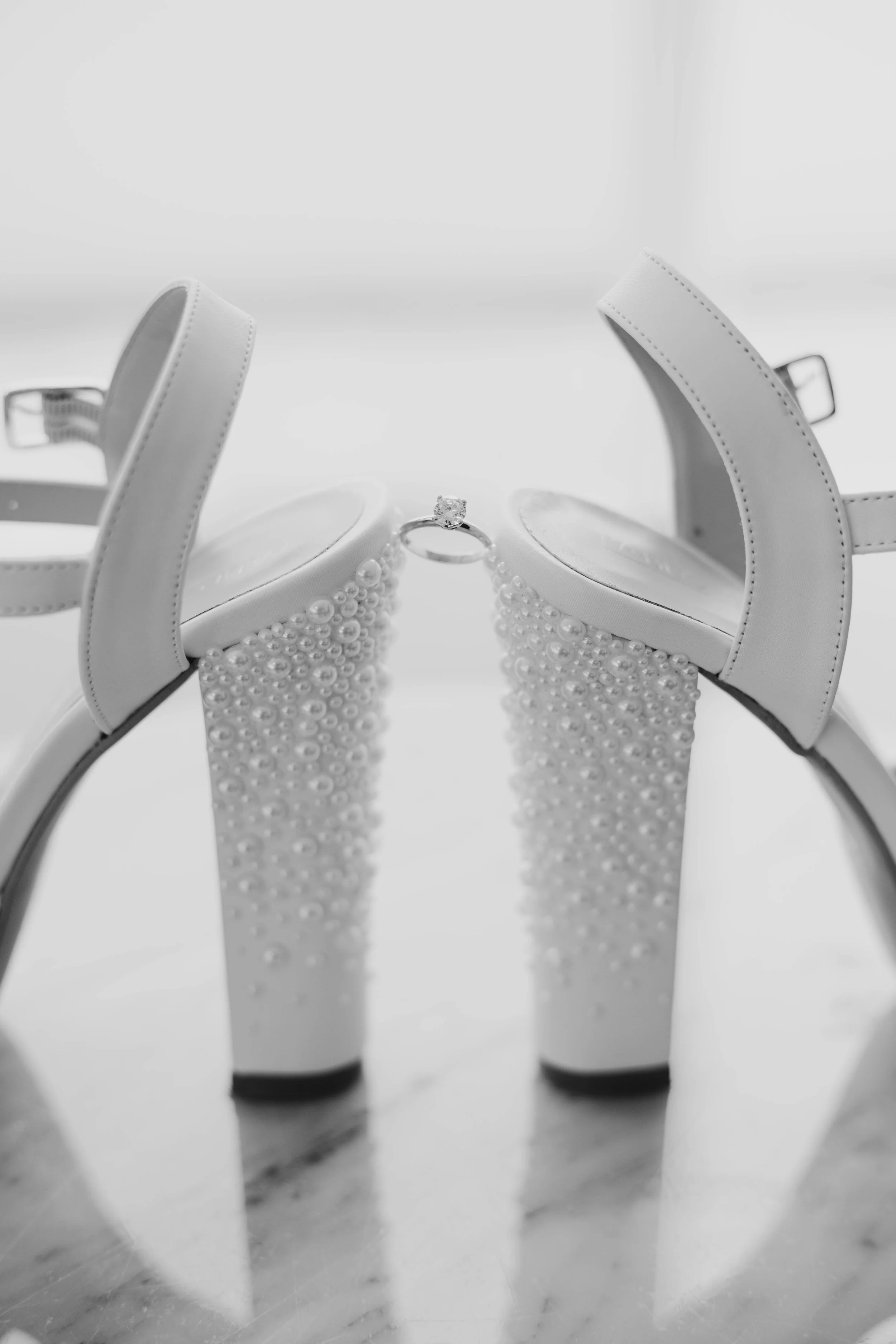 Zapatos blancos de tacón alto con perlas y anillo de compromiso en medio.