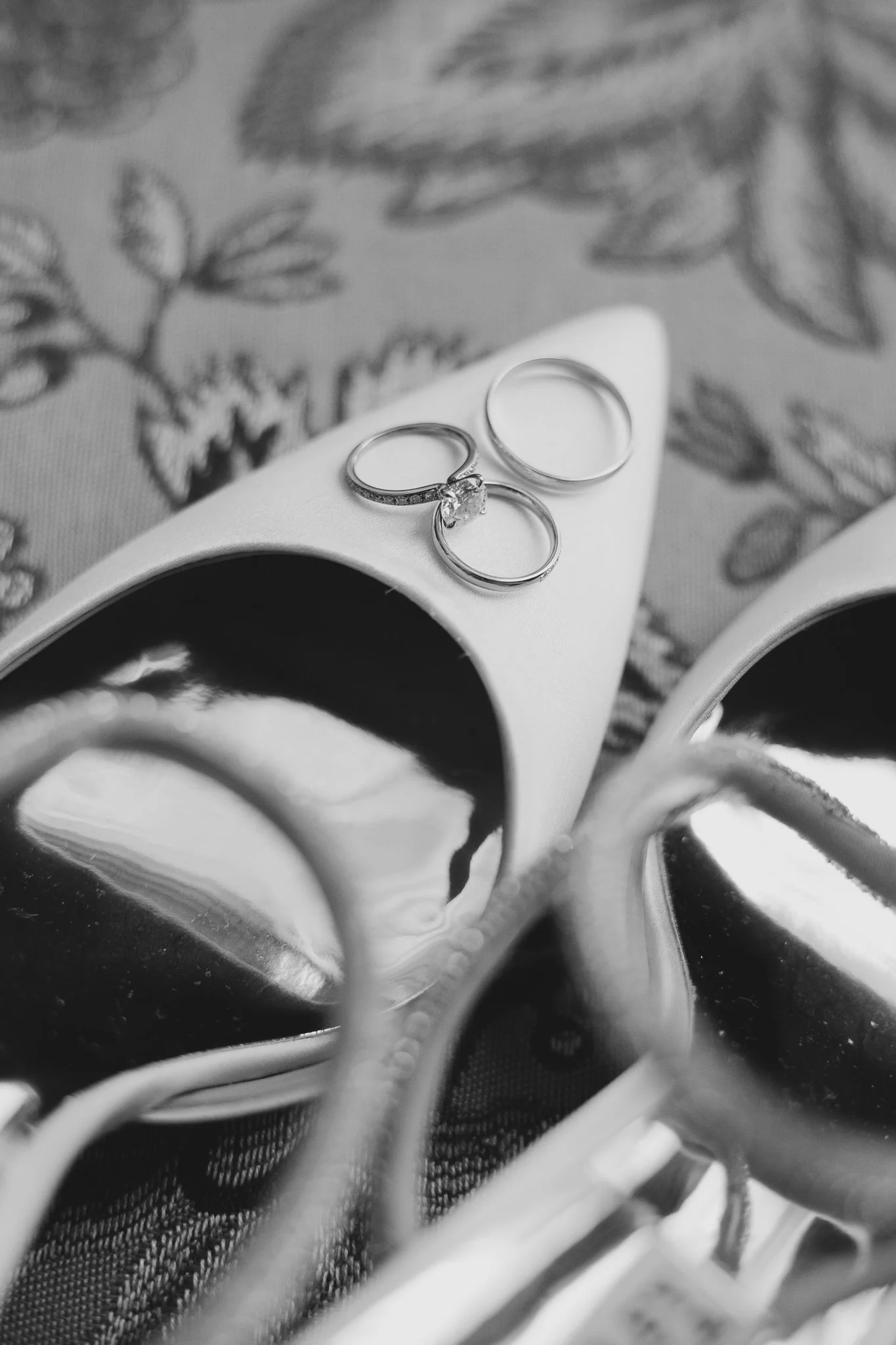 Anillo de compromiso con diamante y dos alianzas en un zapato de tacón alto con estampado floral en el fondo, en blanco y negro.