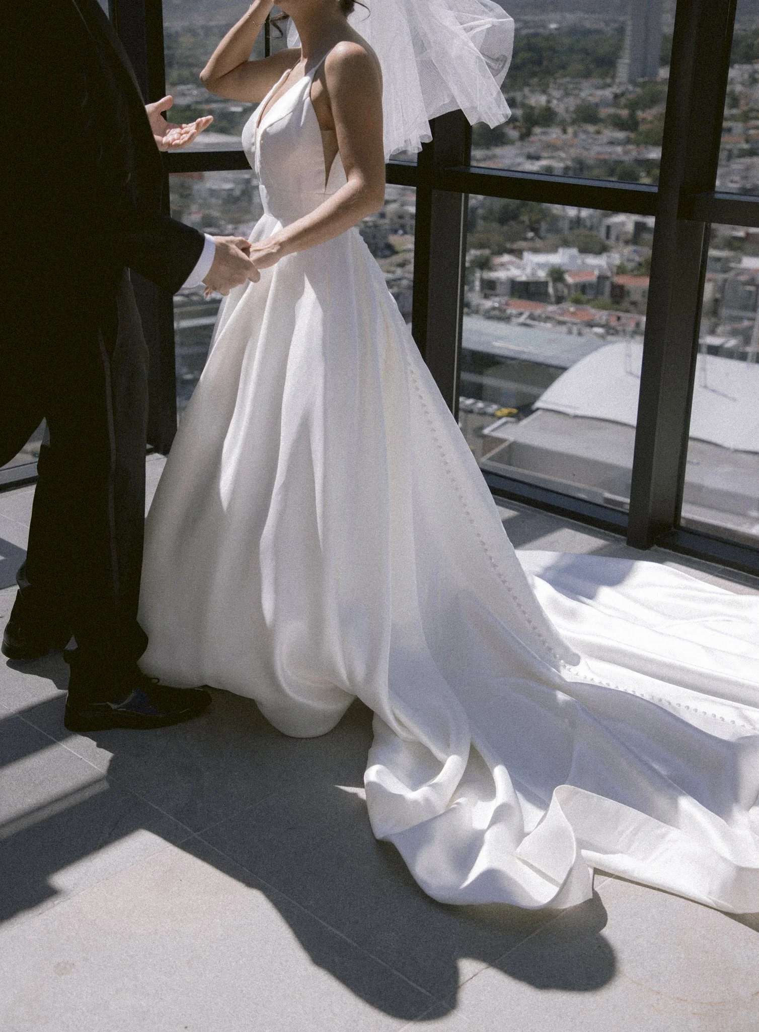 Una pareja en su boda, sosteniéndose las manos, en un lugar con vista panorámica de una ciudad.