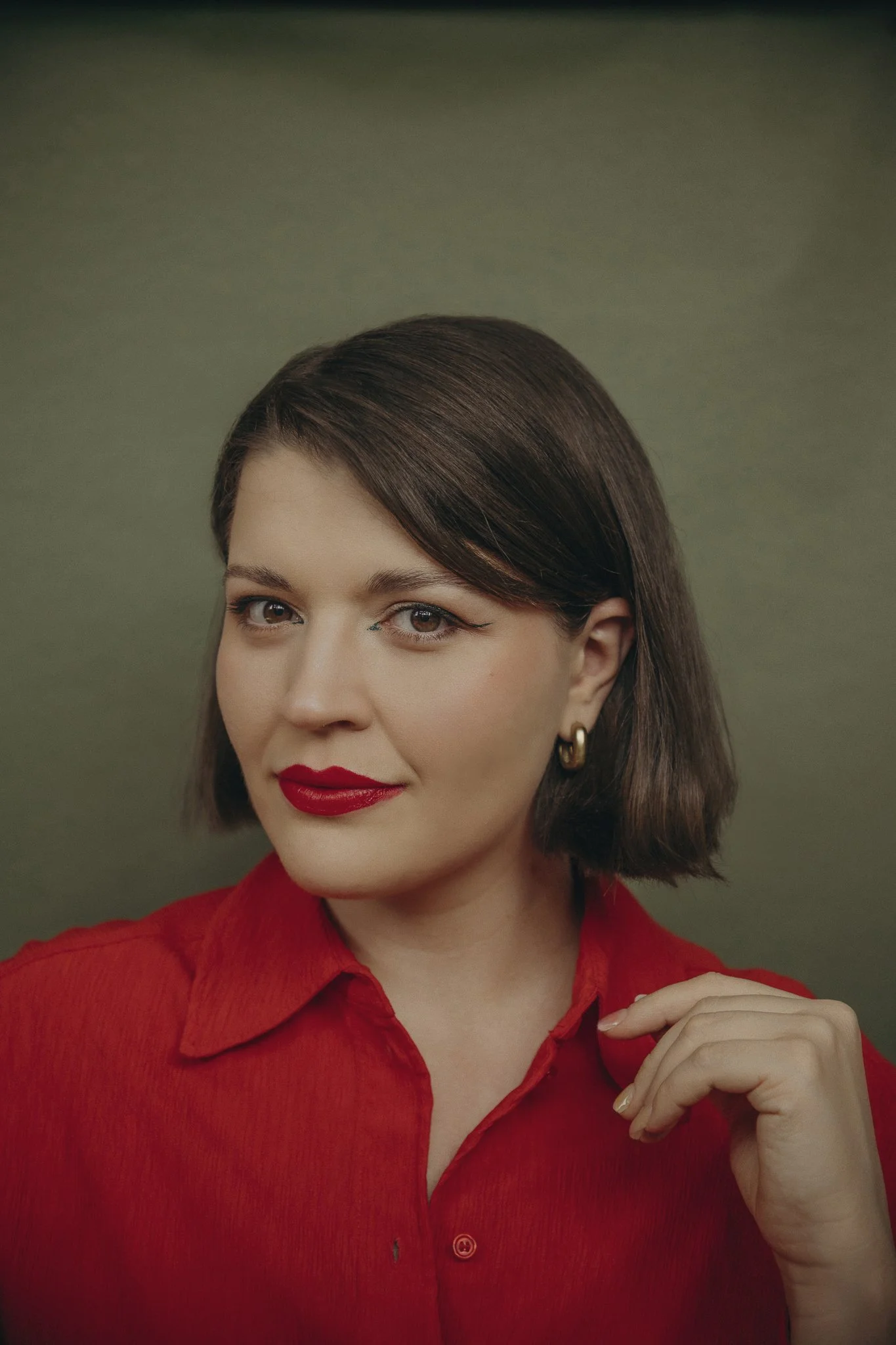 Retrato de una mujer con cabello castaño corto, ojos claros y labios rojos, usando blusa roja y aretes dorados, con fondo verde, Noemi Fotógrafa de Limón Real Photography