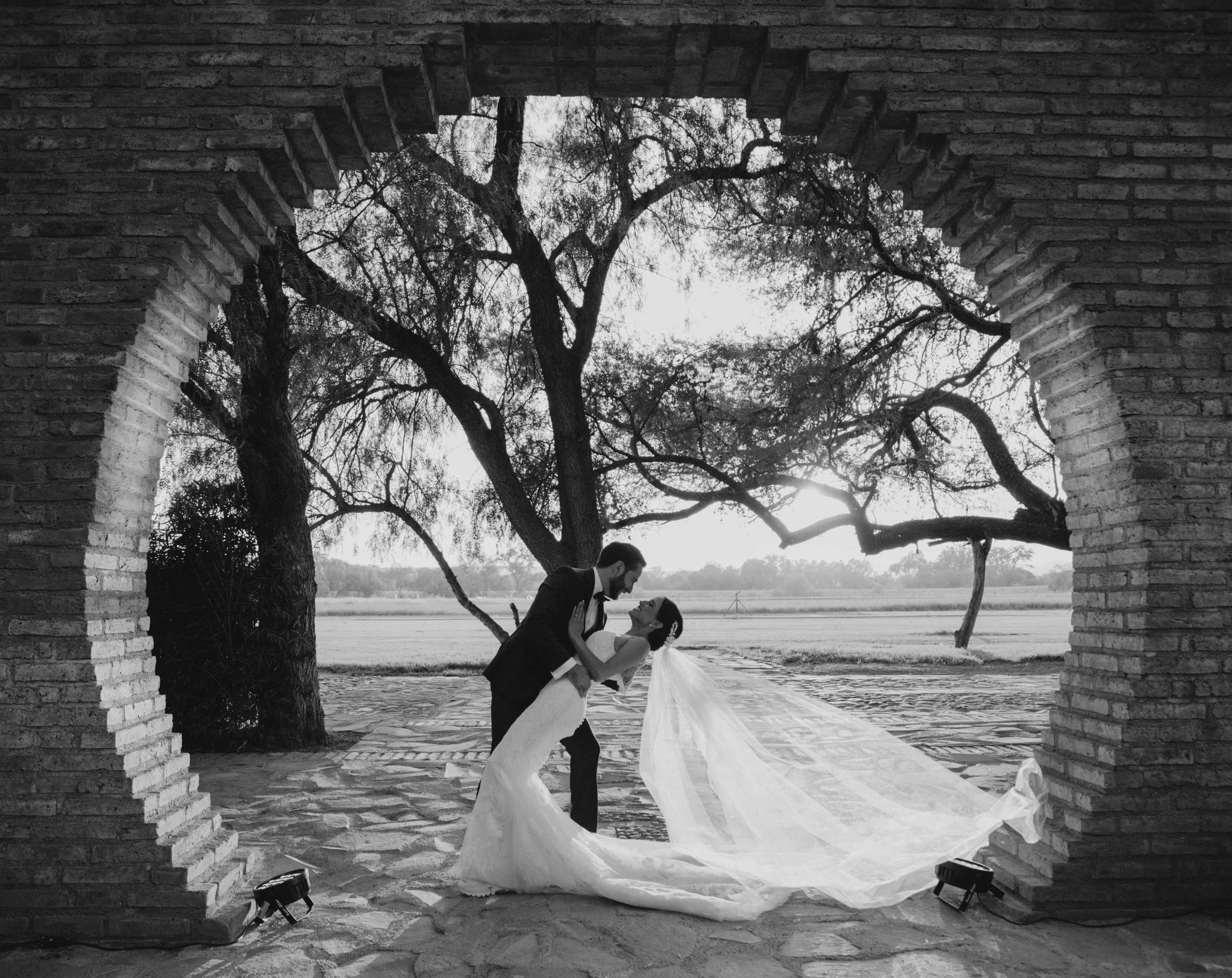 Pareja de novios bailando bajo un árbol en un escenario al aire libre, atrapados en un abrazo, con un arco de ladrillos a su alrededor y luces en el suelo.