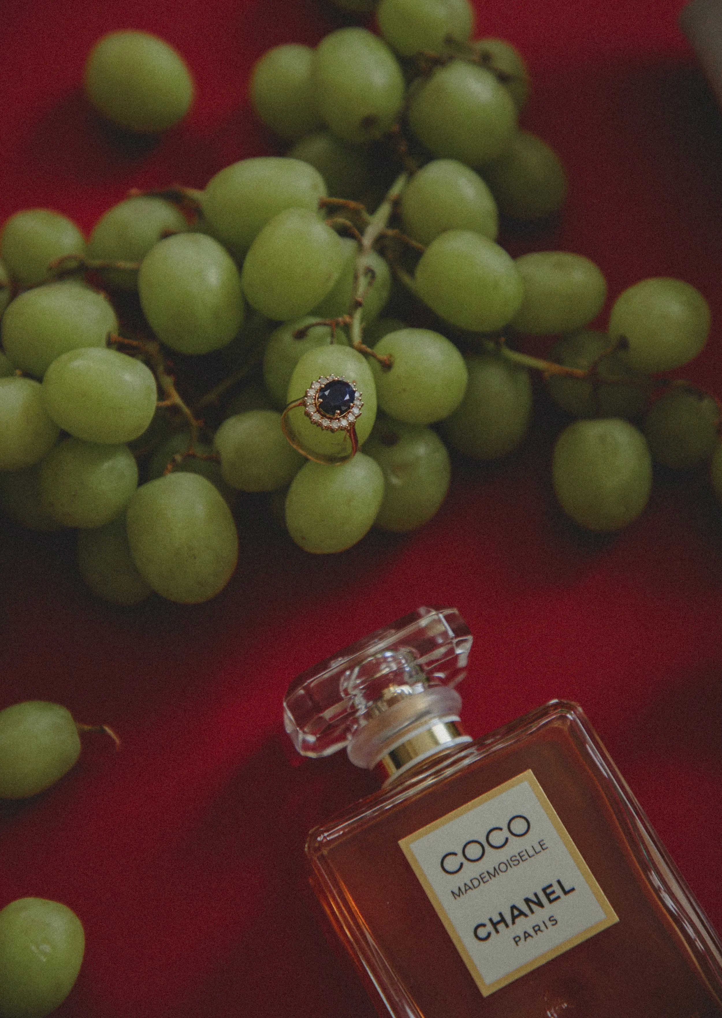 Ramillete de uvas verdes con un anillo de joyería con piedra azul y biseles con diamantes y un frasco de perfume Chanel Coco Mademoiselle sobre un fondo rojo.
