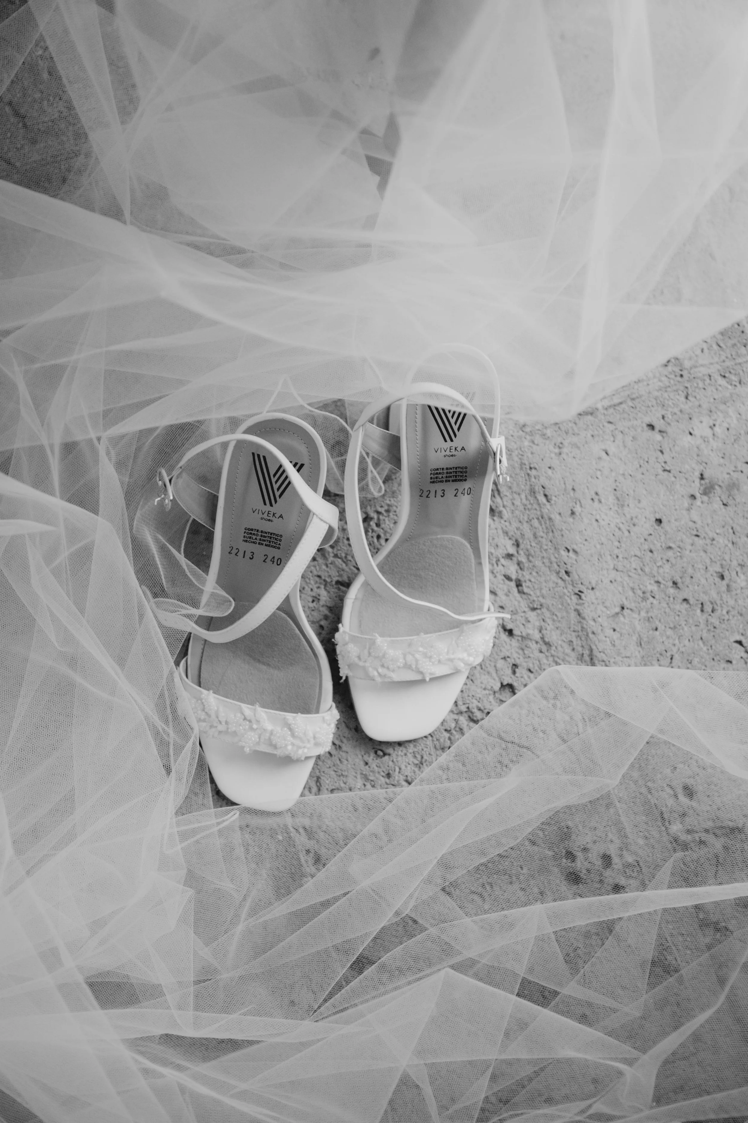 Zapatos de novia blancos con detalles en encaje, sobre una superficie de concreto, rodeados de tela de tul.