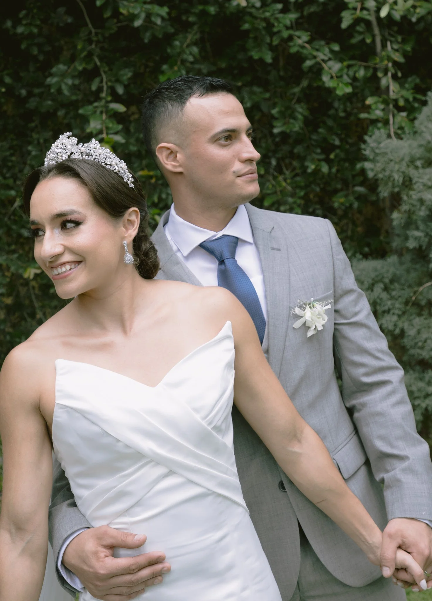 Una pareja de novios en su boda, ella con vestido blanco y tiara, sonriéndo, y él con traje gris y corbata azul, en un fondo de arbustos verdes.