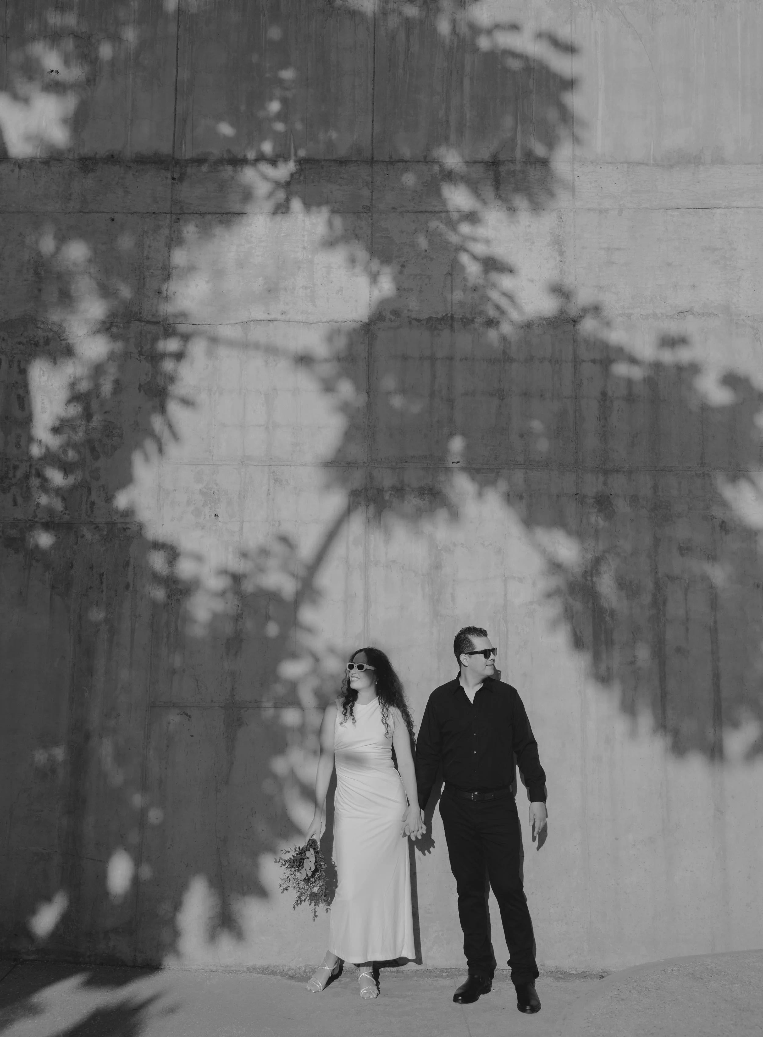 Pareja caminando de la mano frente a un muro de concreto, la mujer viste un vestido largo y sostiene un ramo, y ambos llevan gafas oscuras, en blanco y negro.