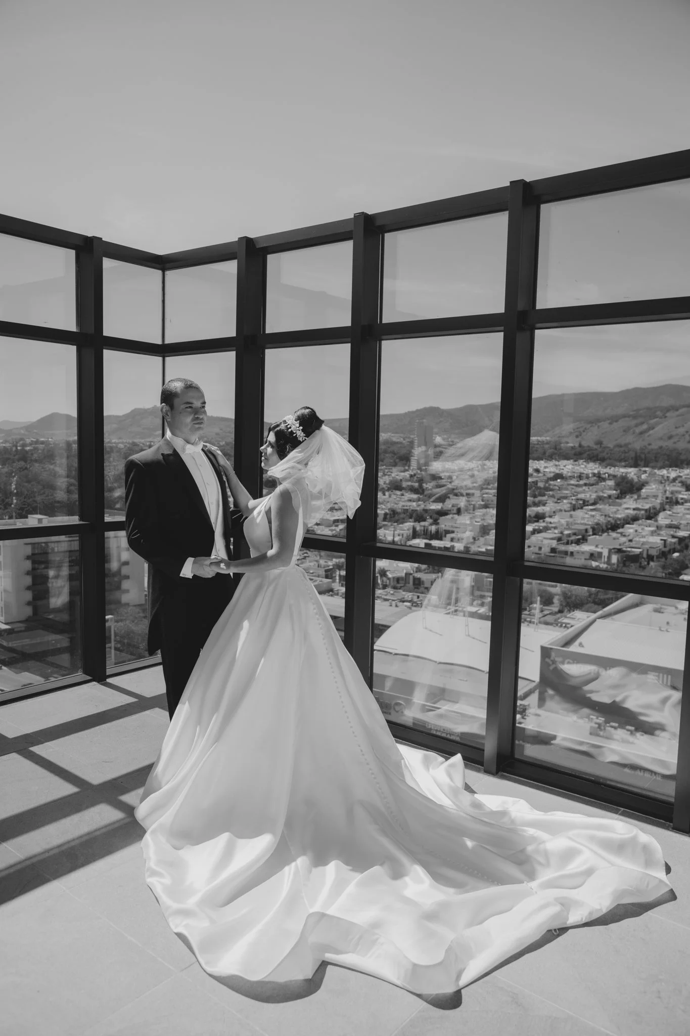 Una pareja de novios en su boda, en un lugar con paredes de vidrio que muestra una vista panorámica de la ciudad y las colinas.