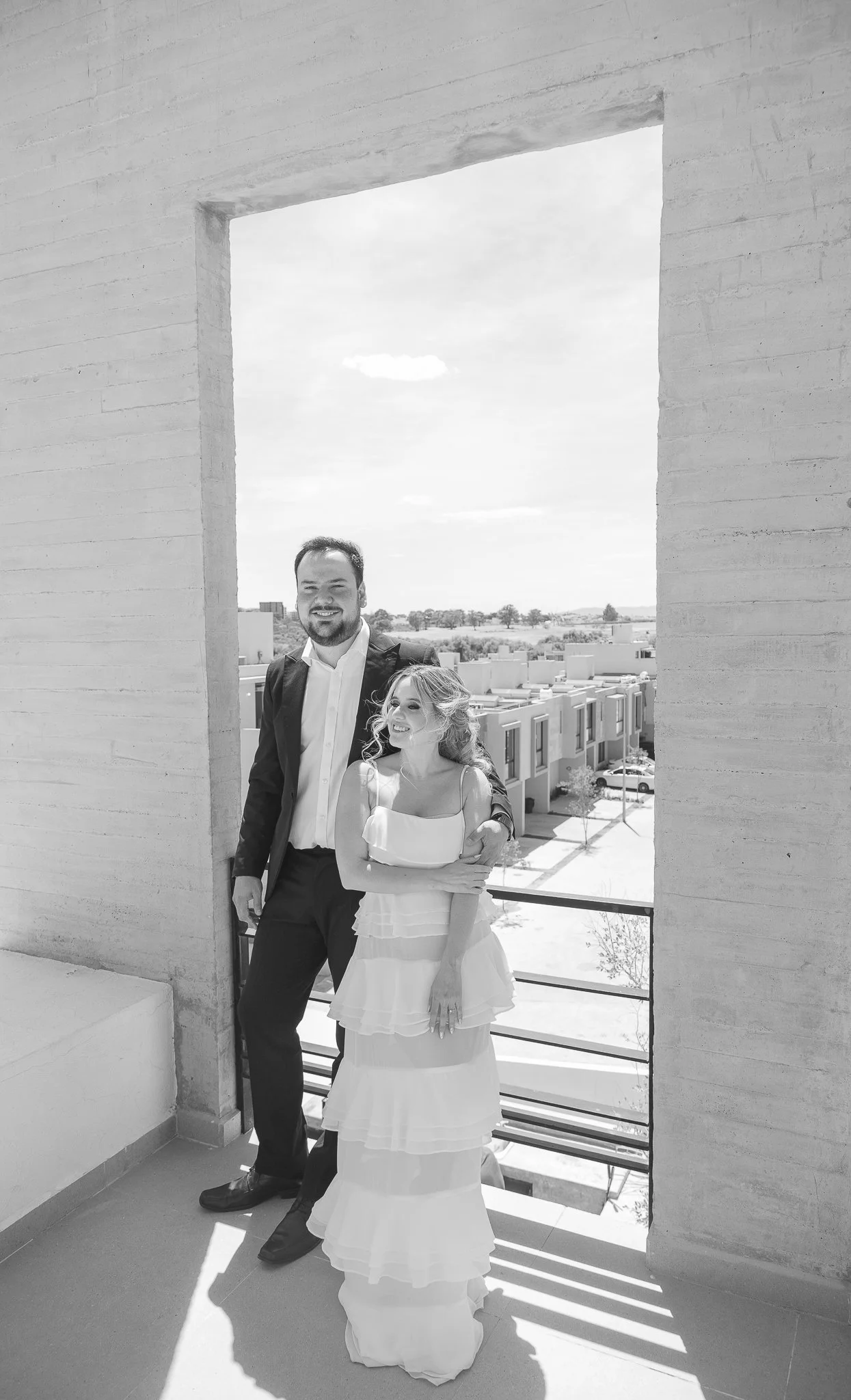 Pareja sonriente en un balcón, la mujer usa vestido largo y el hombre traje.