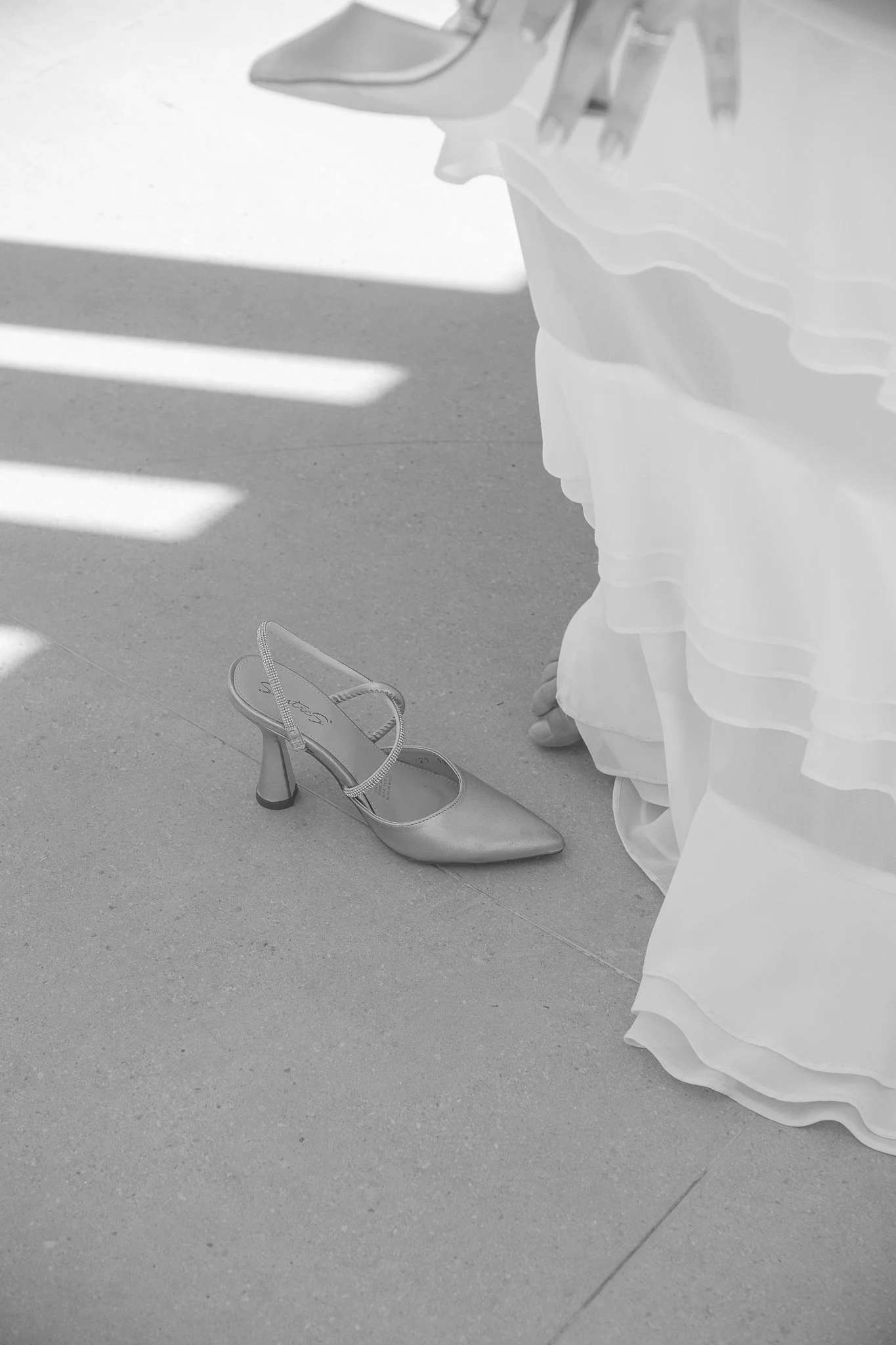 Un zapato de tacón en el suelo junto a una falda larga, en un entorno con sombras en el piso.