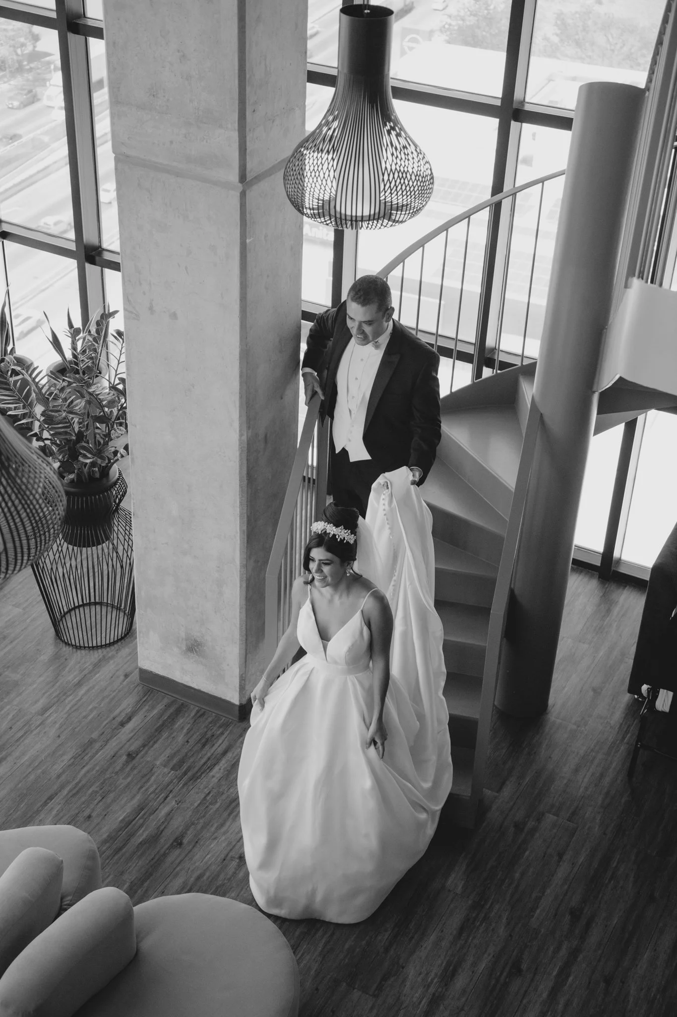 Una novia en vestido de boda bajando una escalera de espiral, con un vestido amplio y una corona de flores en la cabeza, mientras un hombre, probablemente el novio, la acompaña en un entorno moderno con grandes ventanas.