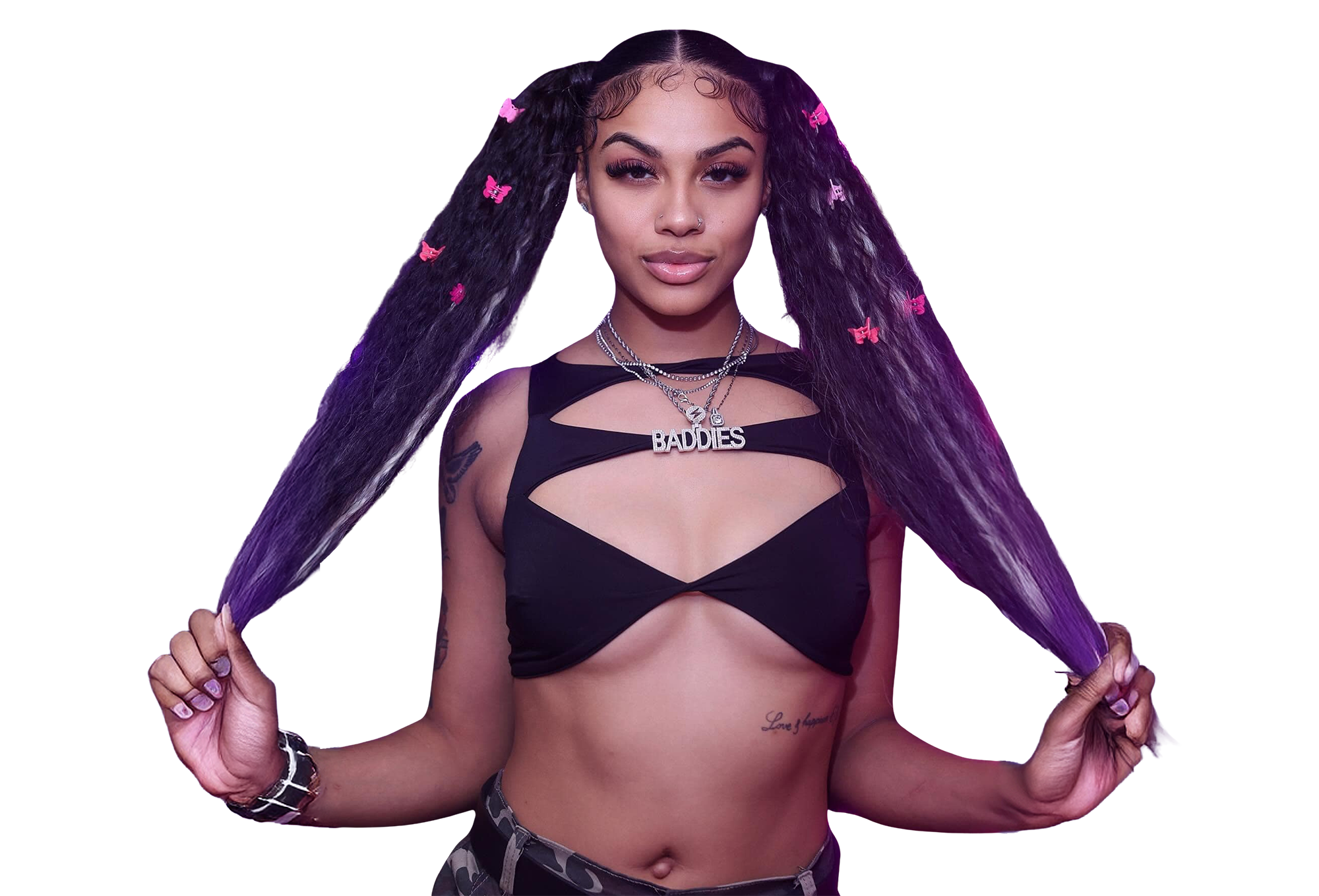 A woman with long, wavy hair with purple and pink butterfly hair clips, wearing a black cutout top, distressed camouflage pants, layered necklaces including one with the word "BAD B.I.D.D.E.S.", and holding her hair.