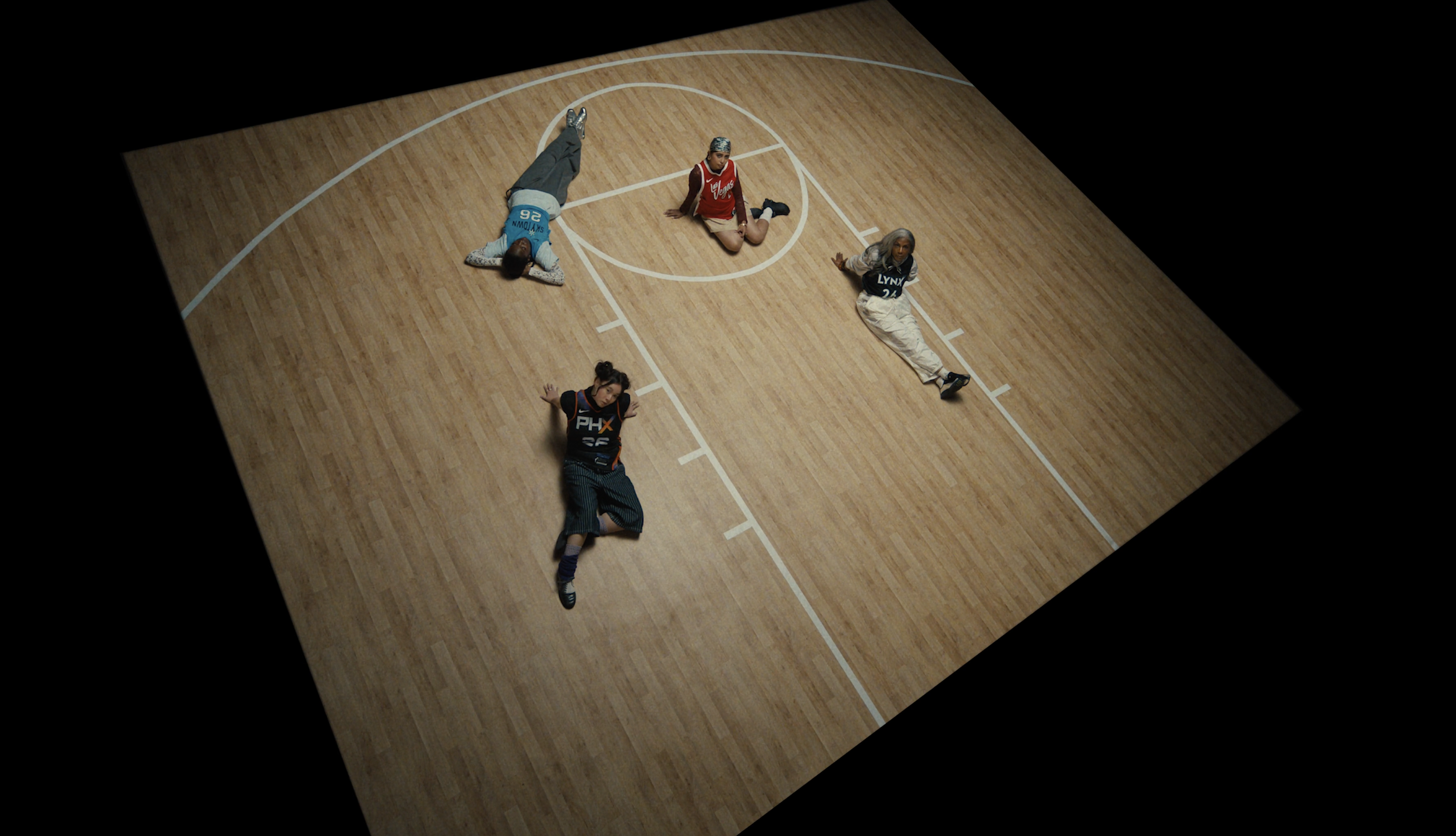 Four people lying on a wooden basketball court, one wearing a Philadelphia Flyers jersey, one in a Los Angeles Lakers jersey, one in a Phoenix Suns jersey, and another person without visible jersey, all in relaxed or resting positions.