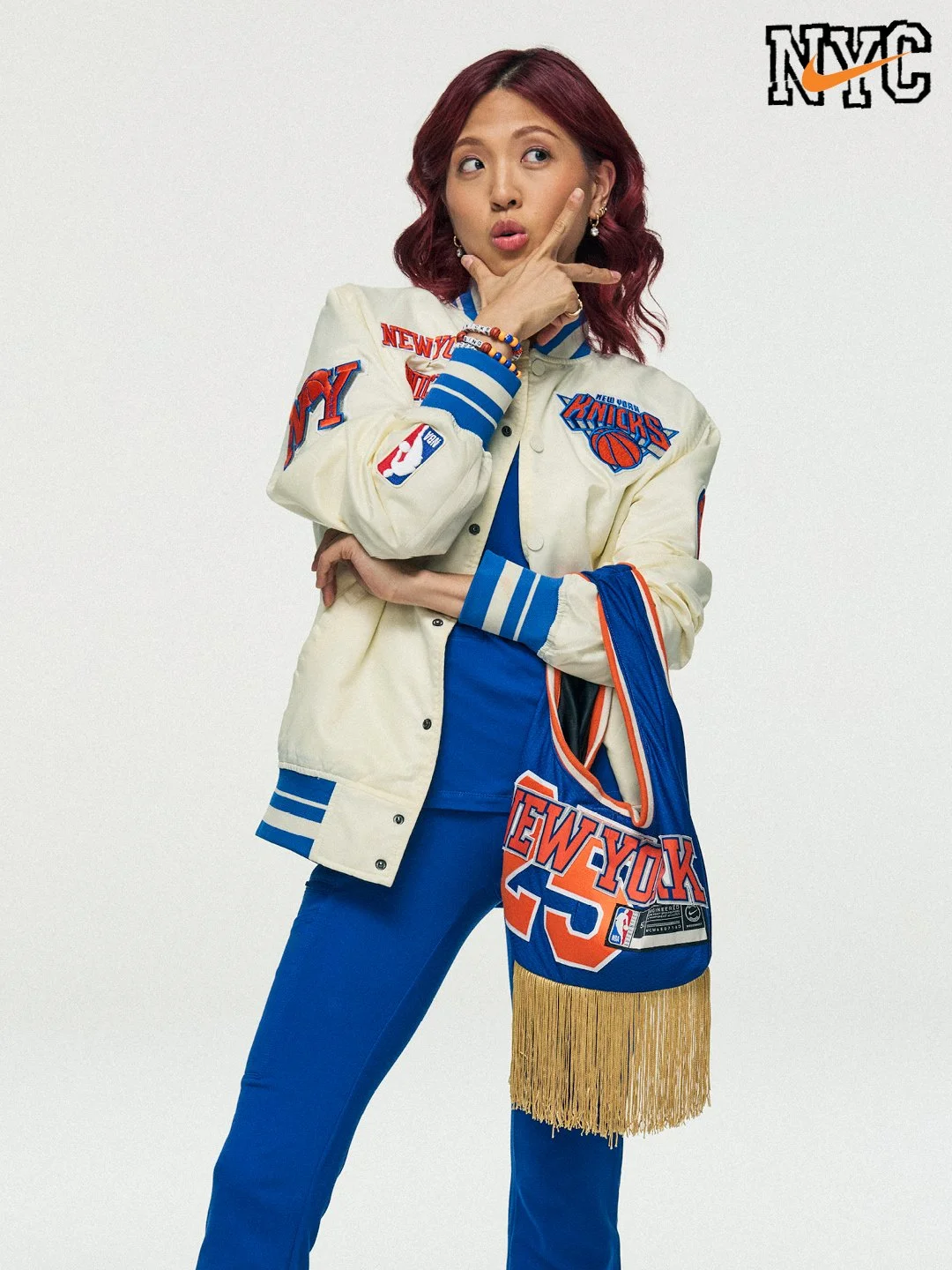 A woman with red hair wearing a New York Knicks varsity jacket, blue pants, and holding a New York Knicks bag with fringe, standing against a plain background.