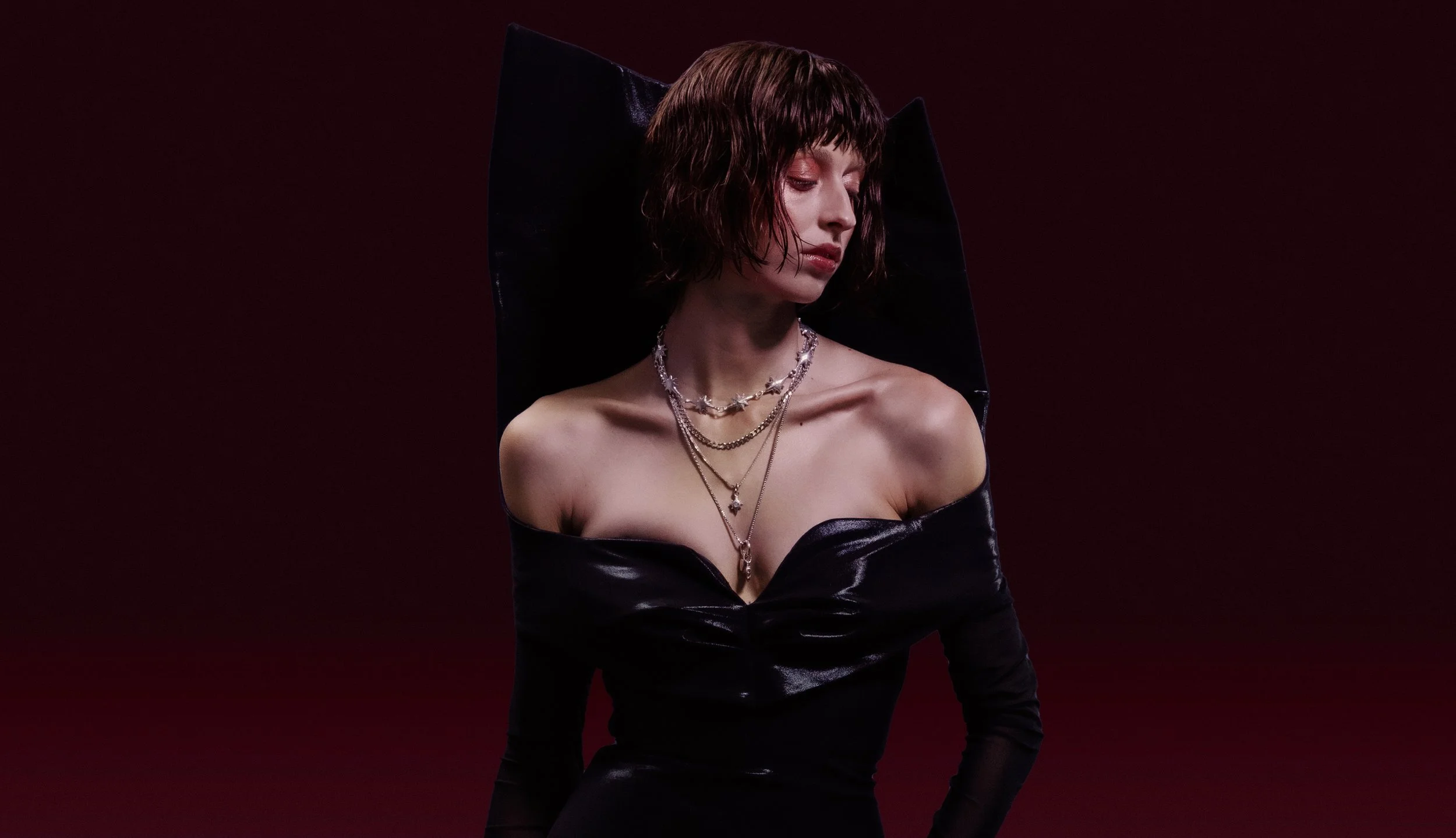 A woman with short brown hair, wearing a black off-the-shoulder dress and multiple layered necklaces, posing against a dark burgundy background.