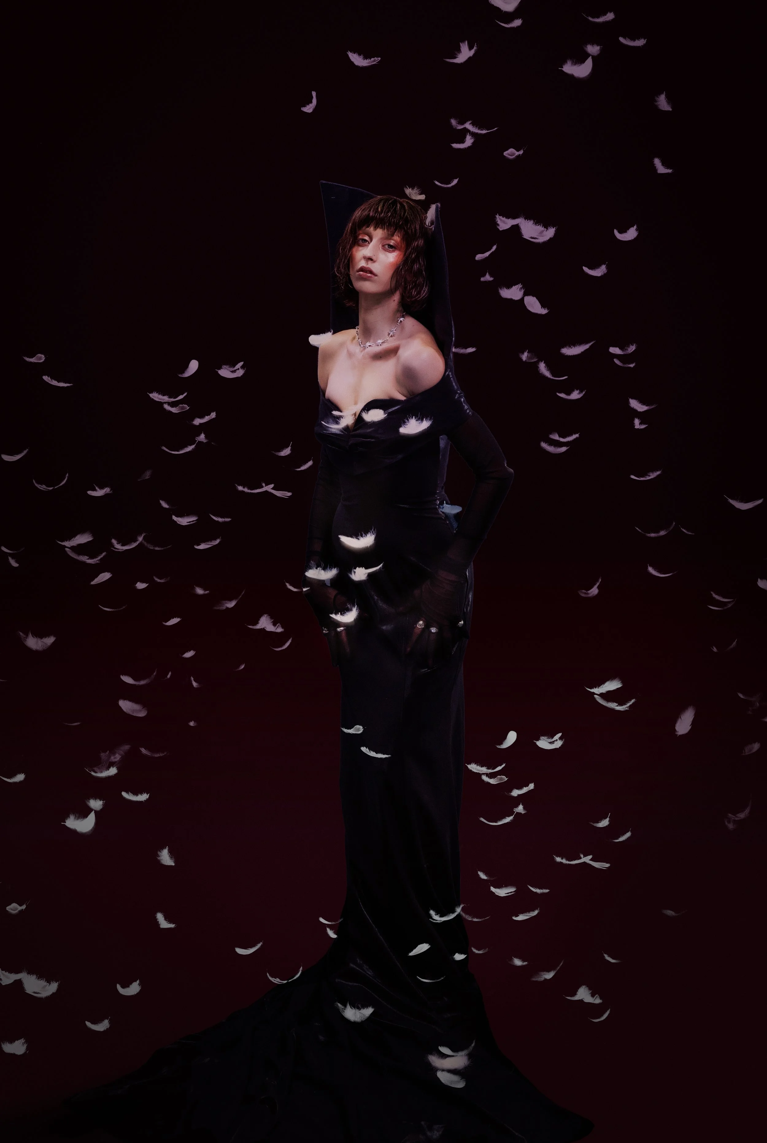 A woman with dark hair and a black outfit standing against a dark background with white feathers floating around her.
