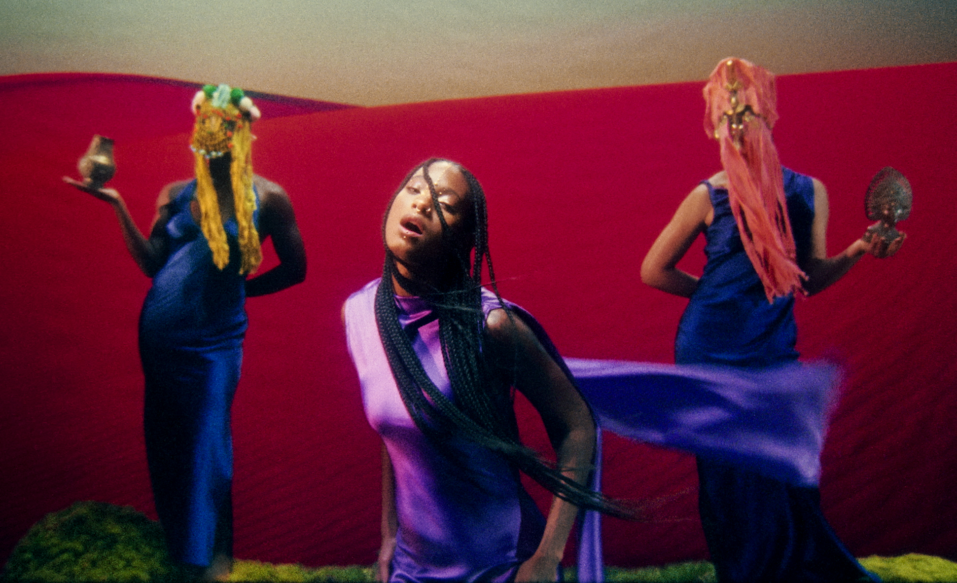 Three women in colorful dresses perform traditional dance with scarves against a red backdrop.