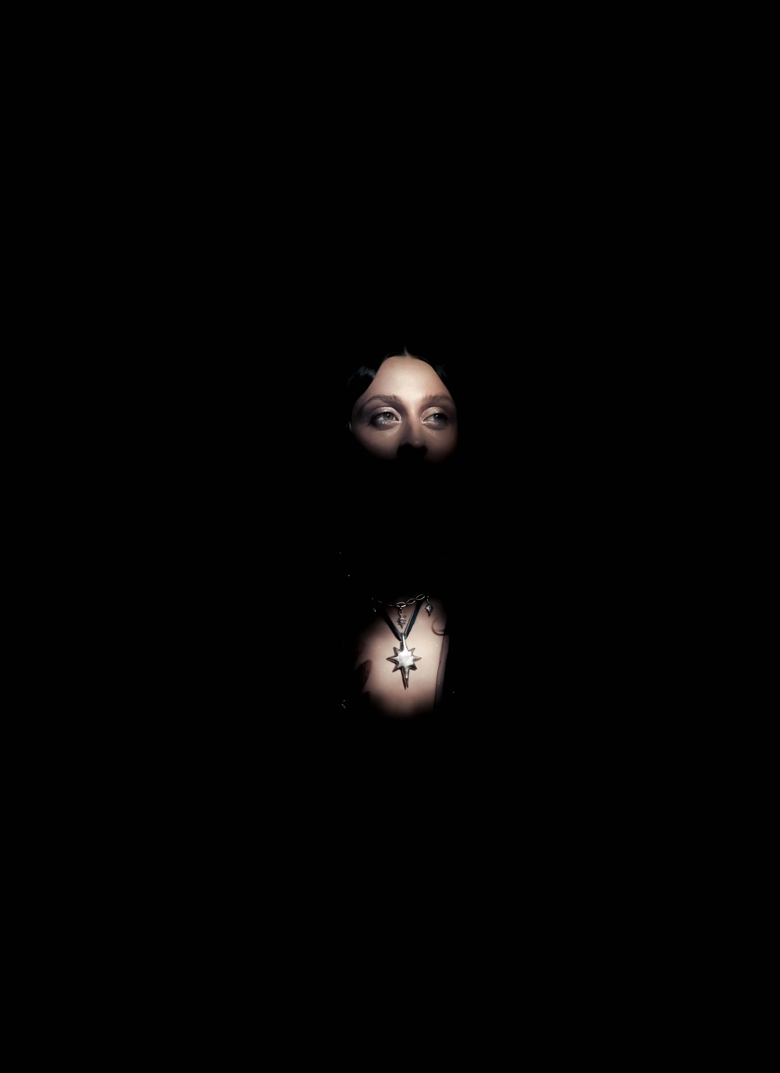 A woman with makeup and jewelry, including a large star-shaped necklace, is illuminated against a dark background, with only her face and upper chest visible.