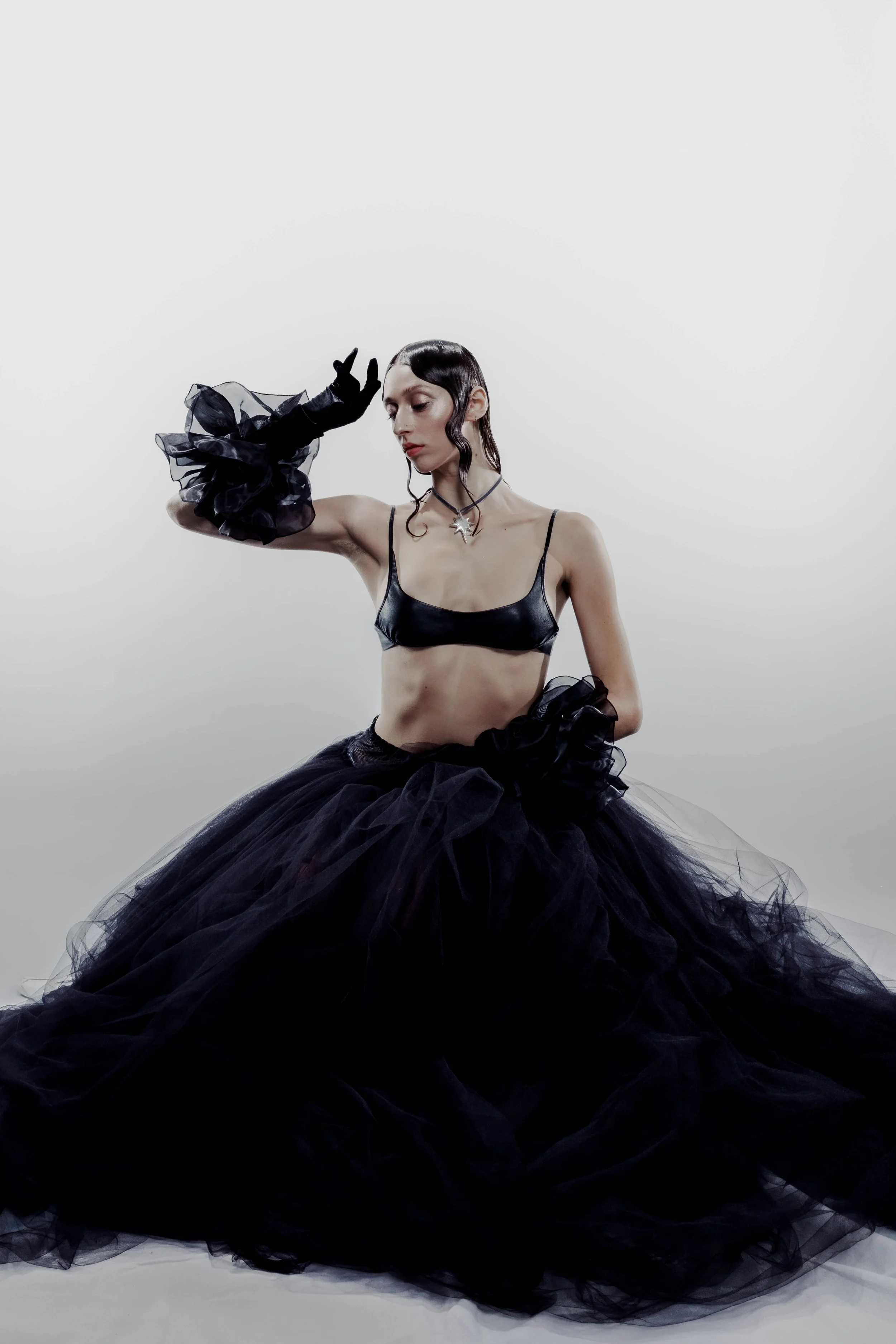 A woman with wet black hair and pale skin wears a black satin bralette, black tulle ruffled layers, and a star-shaped necklace, posing with one arm bent and her hand resting on her head against a plain light gray background.
